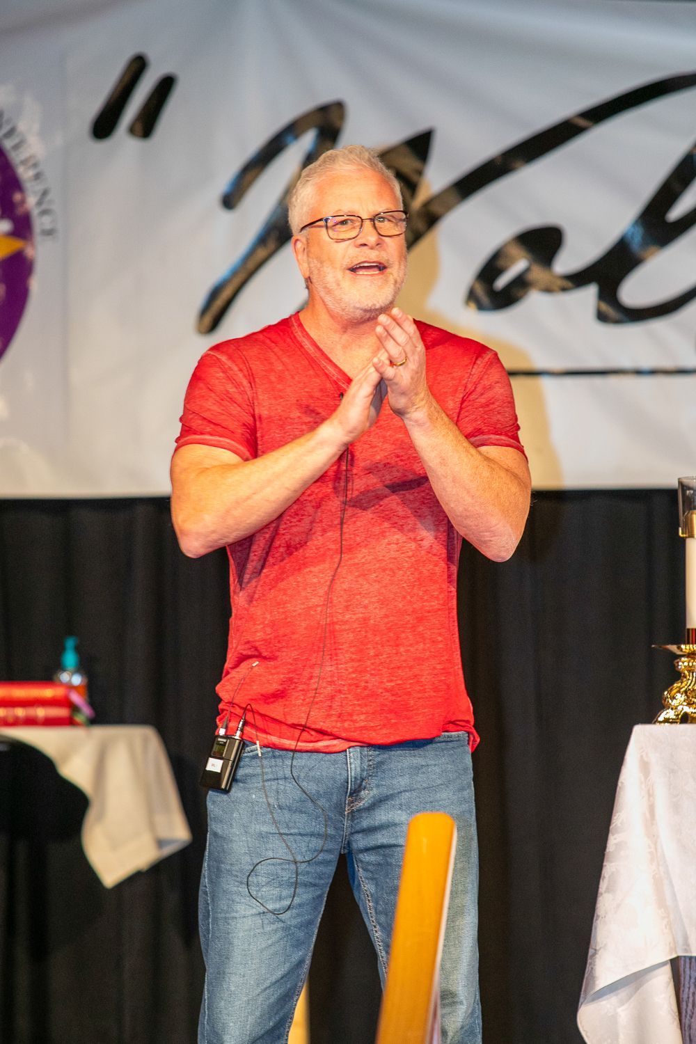 Sean Forrest in red shirt and jeans speaking on stage with hands clasped, in front of a banner and table.
