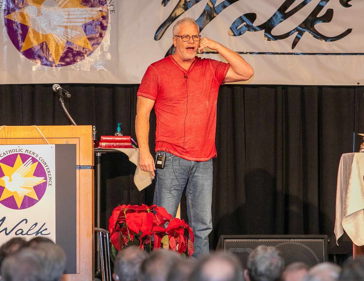 Man in red shirt on stage, hand to forehead, speaking at podium with floral arrangement nearby.