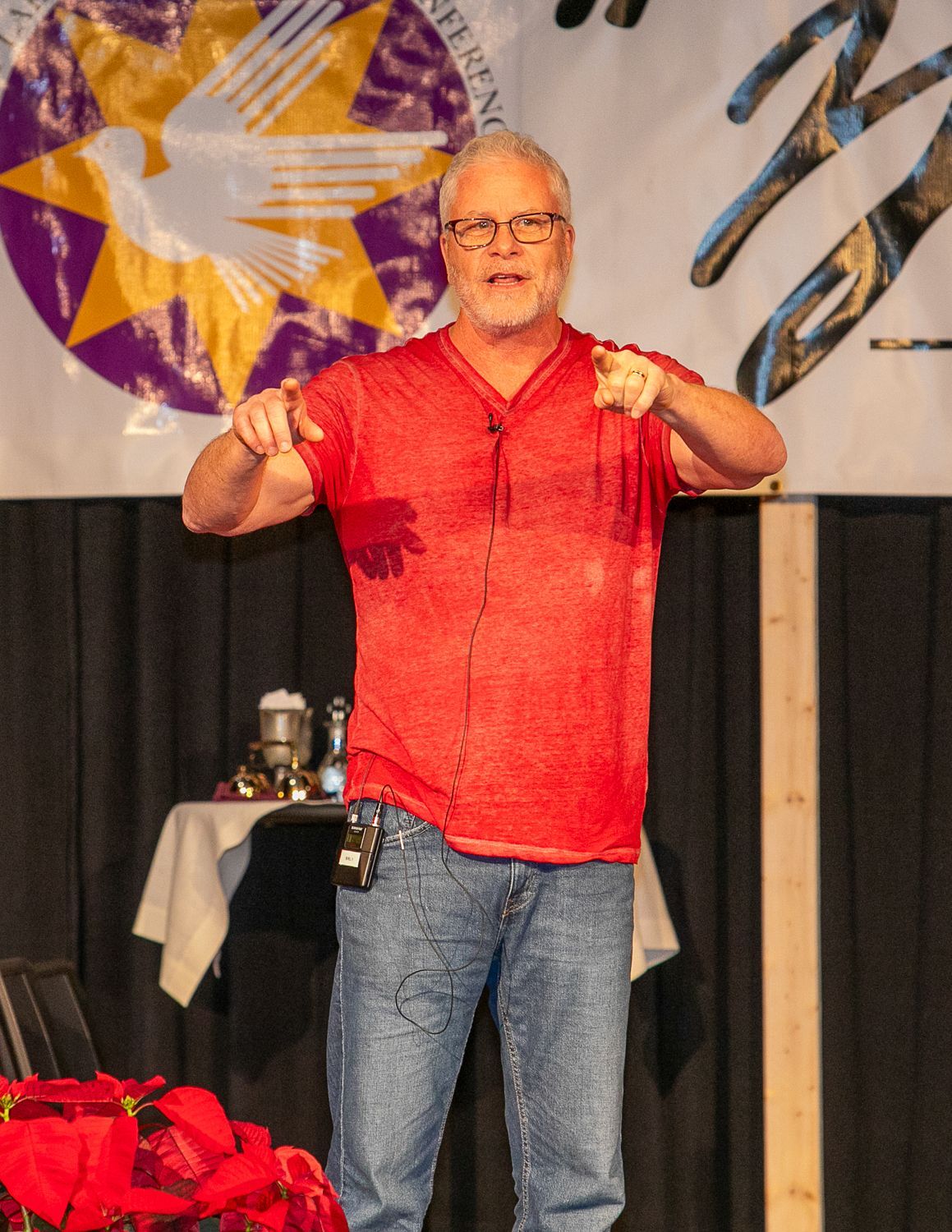Sean Forrest in red shirt and jeans gesturing on stage with abstract backdrop and table behind him