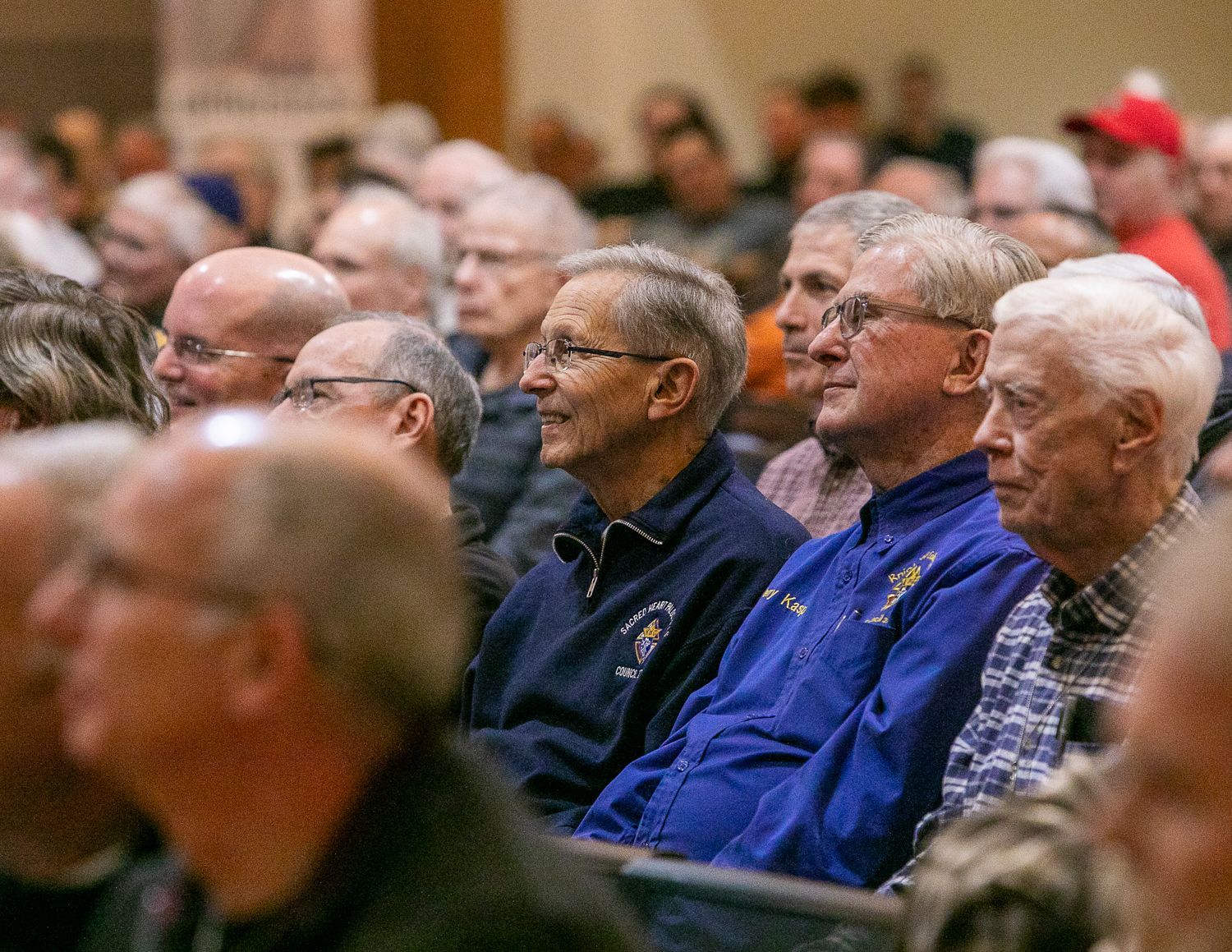 Audience seated in an auditorium, listening attentively, with several people in blue shirts in the front rows