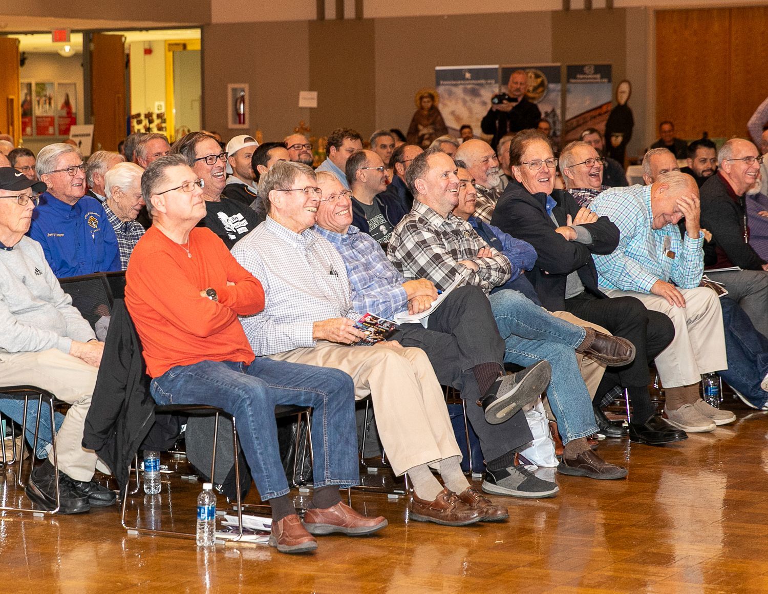 Audience seated in a hall, listening to a presentation, with a man in a red shirt in the foreground.
