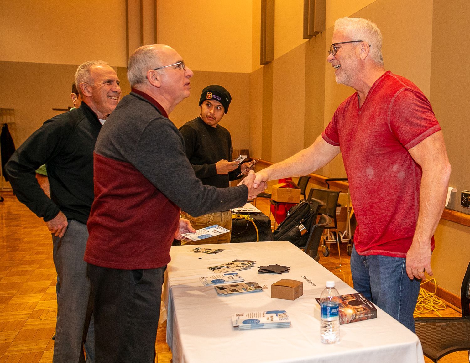 Two men shaking hands across a table at an indoor event, with brochures and items displayed.