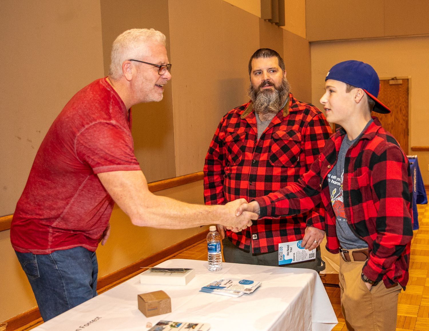 Sean Forrest in red polo shakes hands with man in red plaid at a table; another man stands behind them.