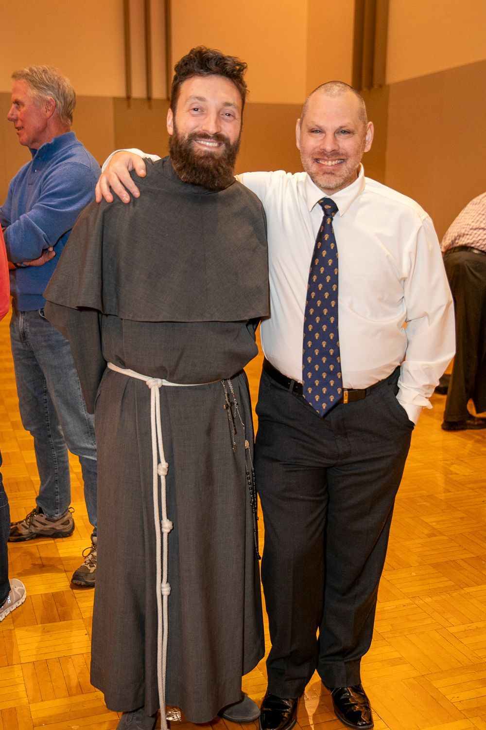 Two men smiling and posing indoors, one in a brown monk’s habit and the other in a white shirt and tie.