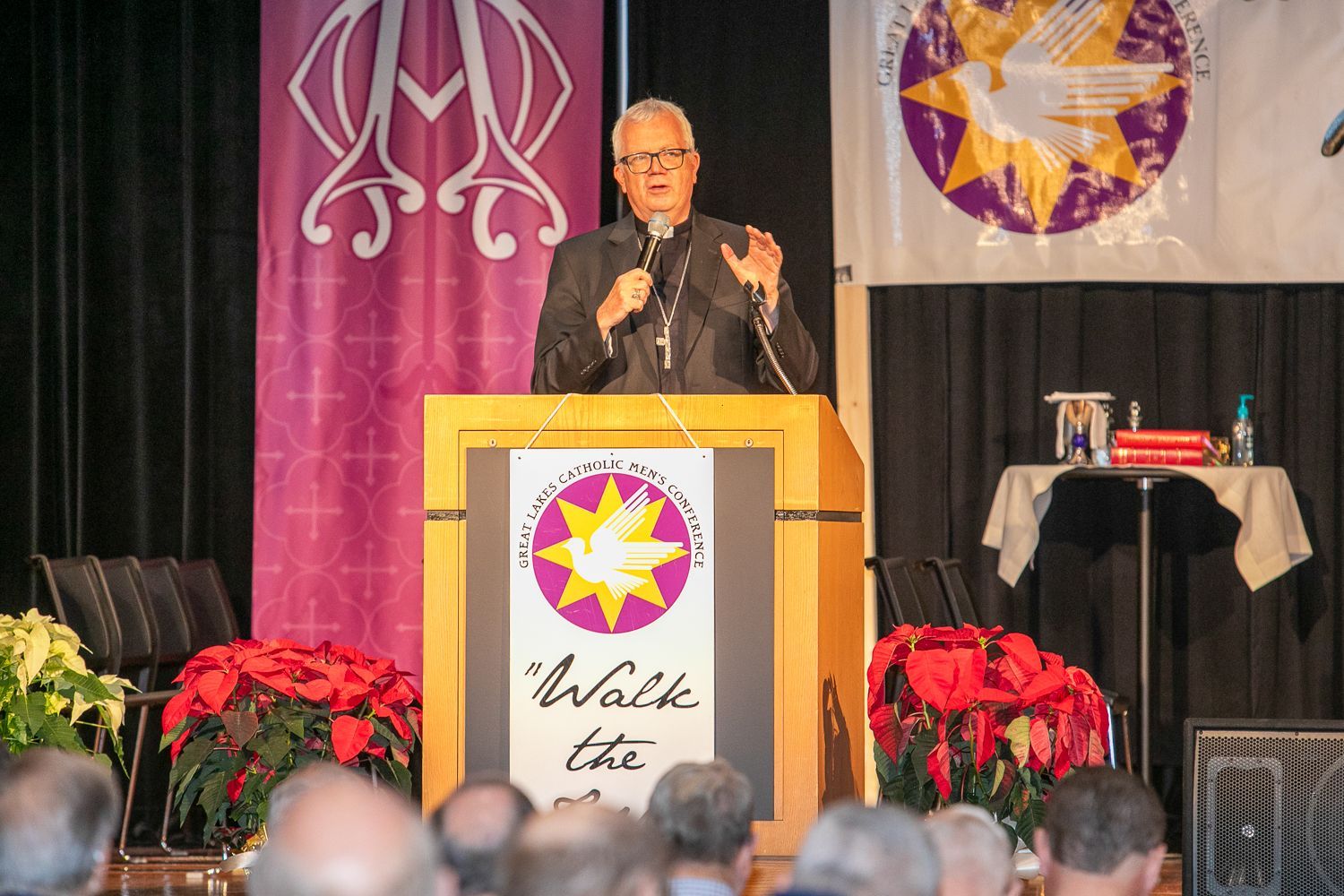 Bishop Hying at podium on stage with banners, addressing audience amid potted poinsettias