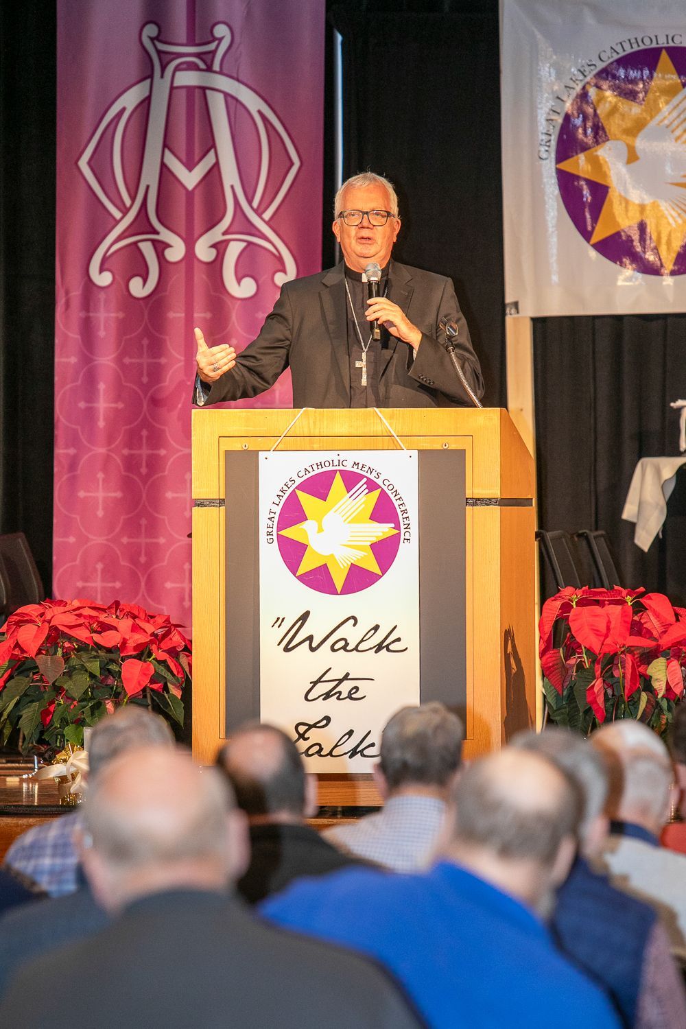 Bishop Hying at the podium addressing the audience during an indoor event, with banners and poinsettias behind him.