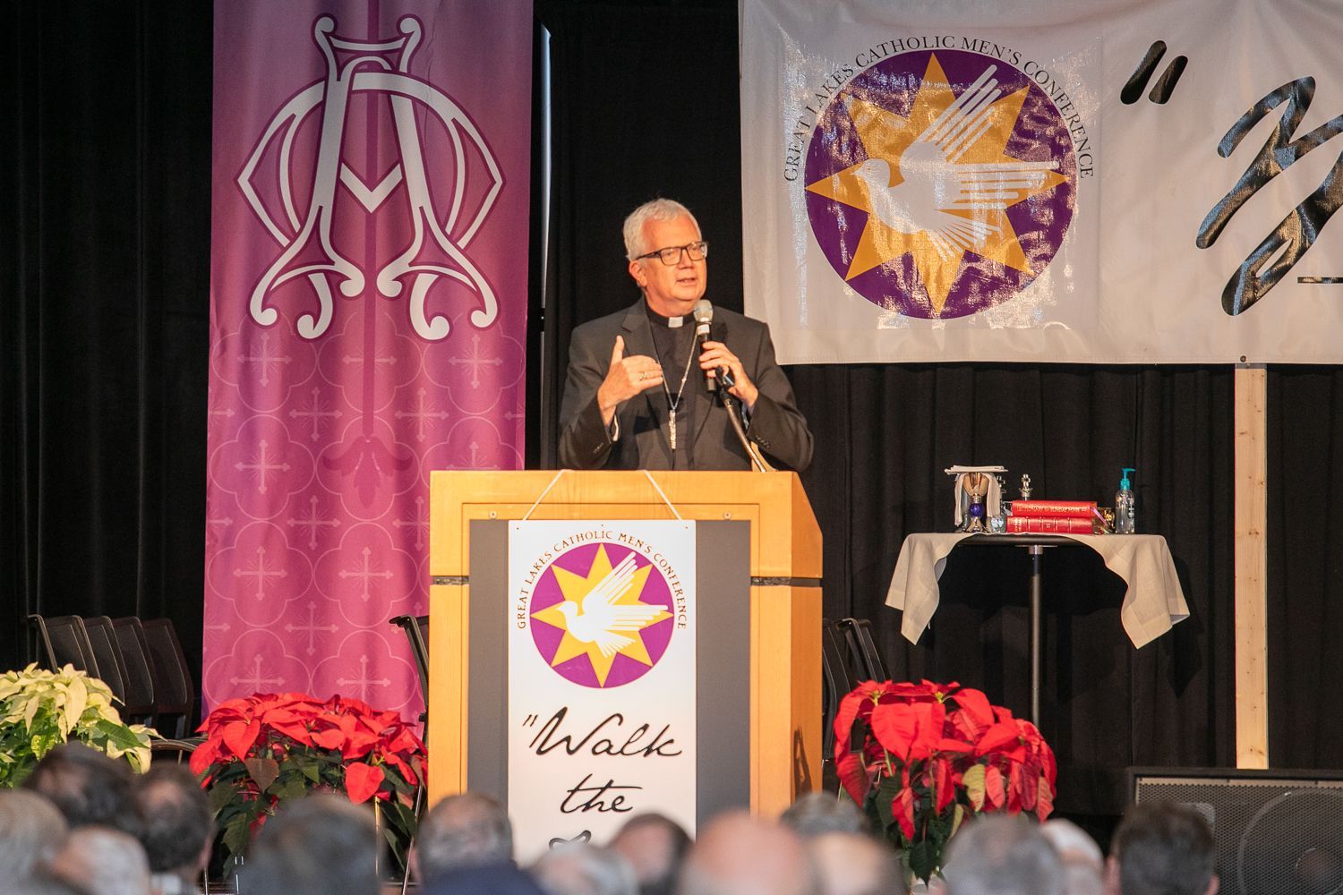 Speaker at podium on stage with purple and gold banners, addressing an audience at a formal event