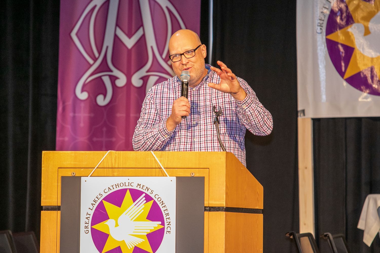 Man speaking into a microphone behind a wooden podium on a stage with banners