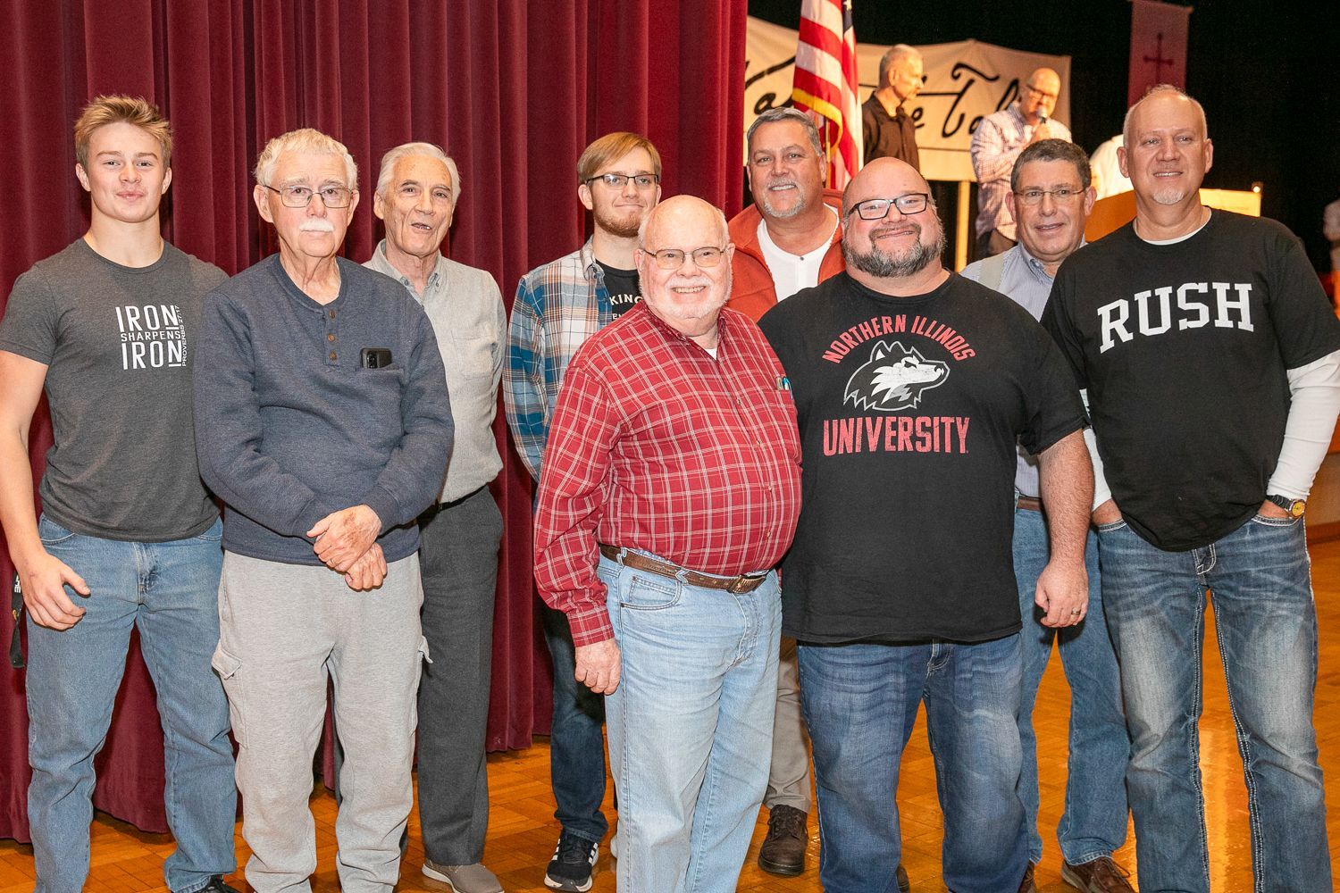 Eight men standing on a stage in front of red curtains, posing for a group photo.