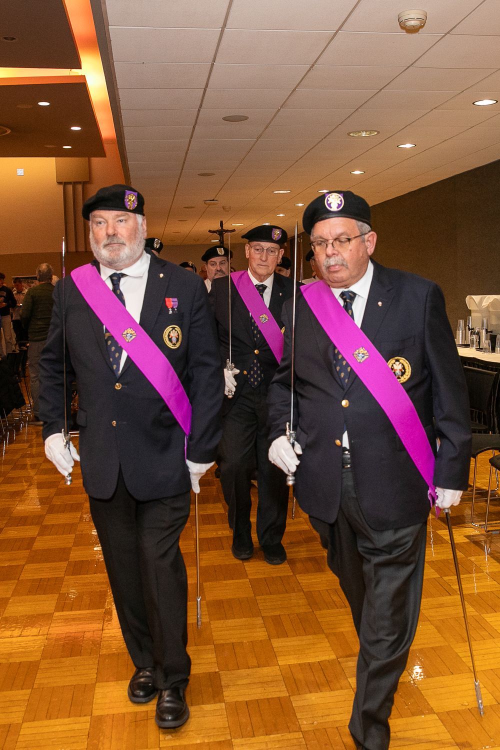 Two uniformed men wearing magenta sashes march indoors on a parquet floor, holding canes.