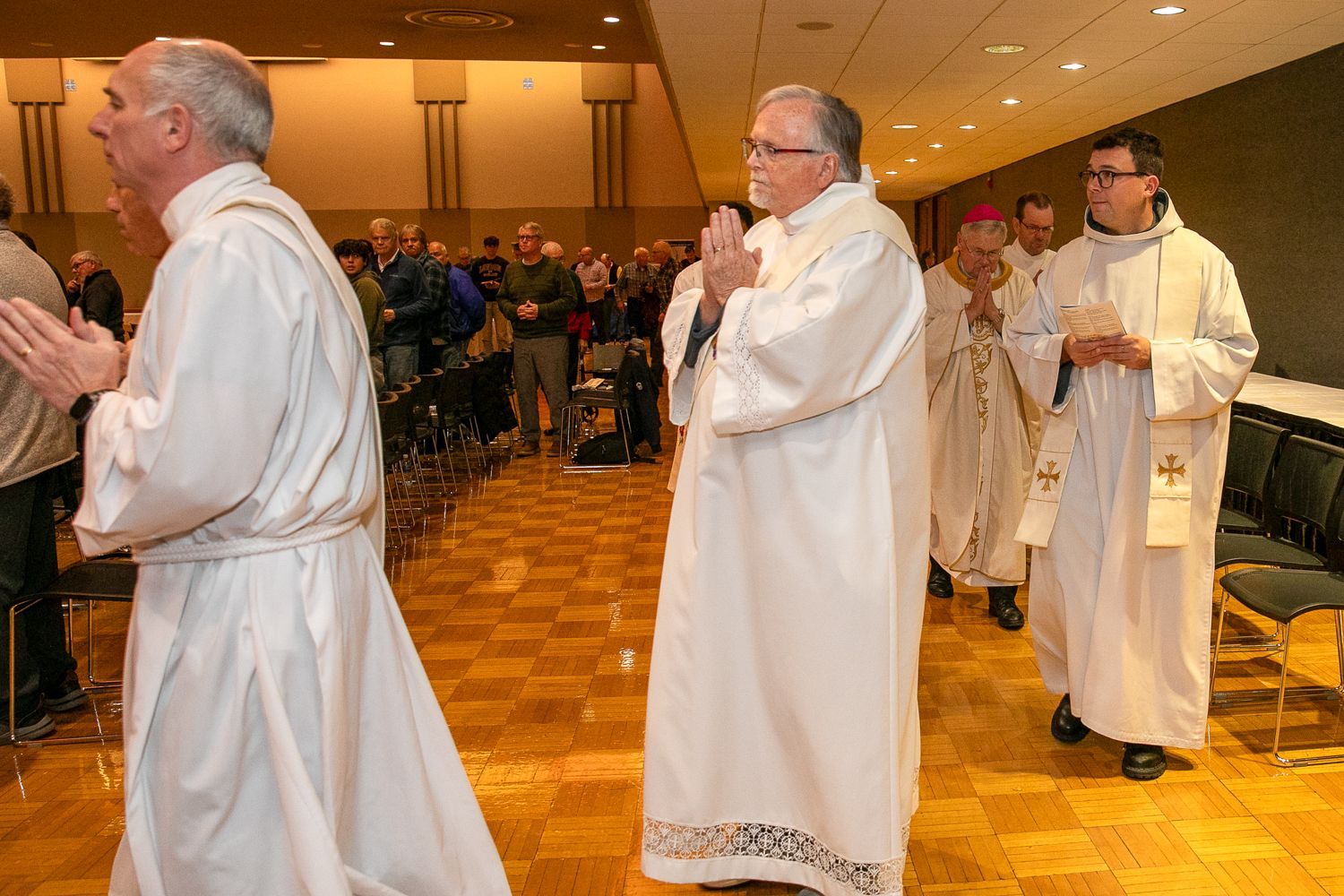 Clergy in white robes processing through a church aisle during a service, with congregants seated in the background