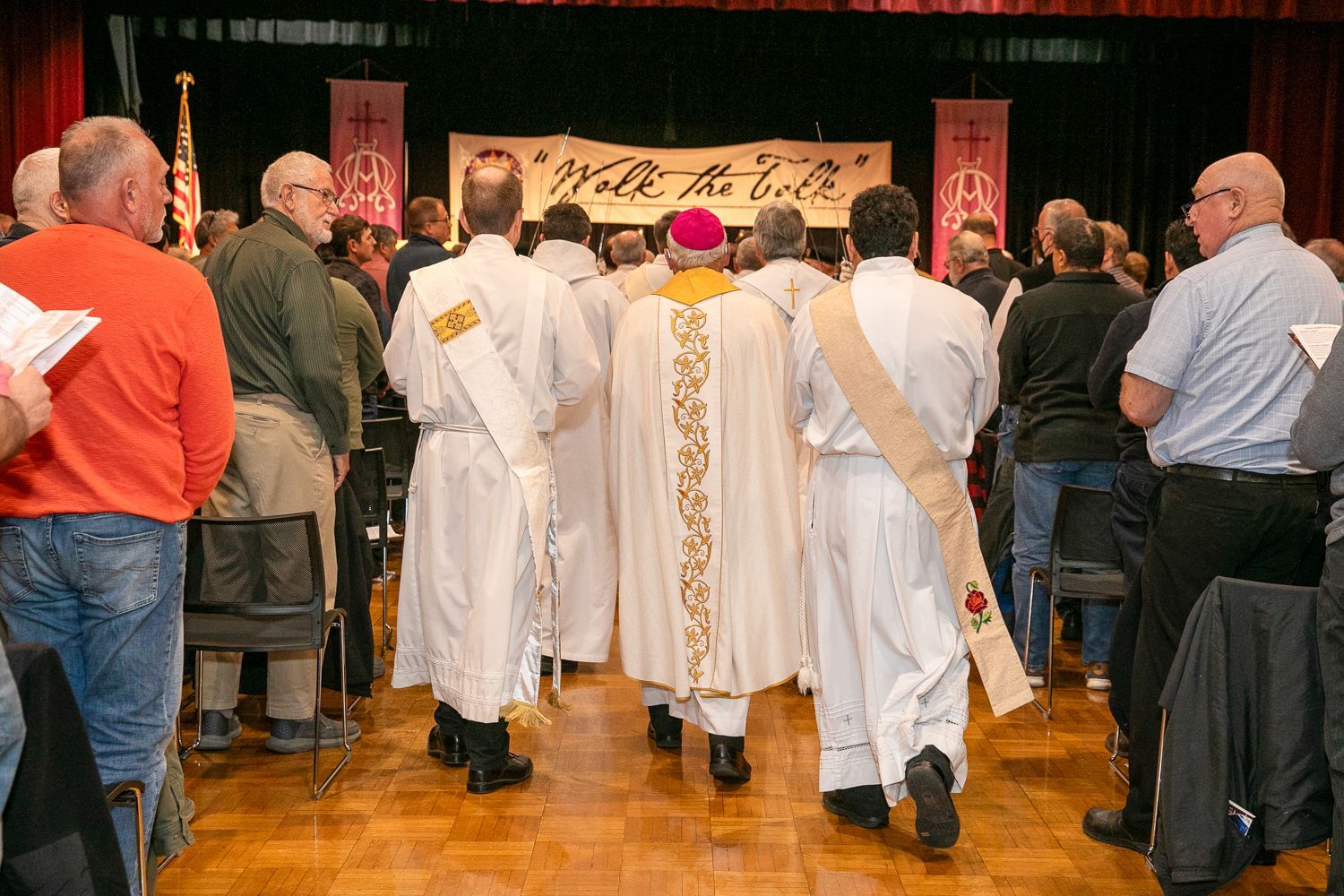 People in white robes walking through a crowded indoor venue toward a stage or booth