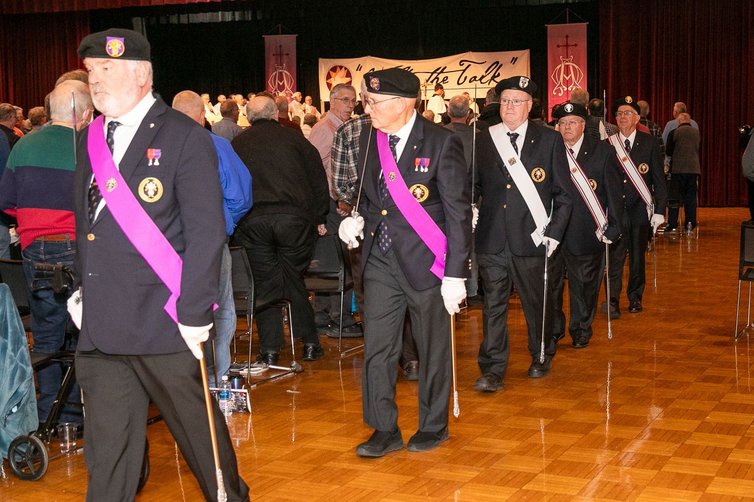 Uniformed veterans marching indoors with purple sashes during a formal ceremony