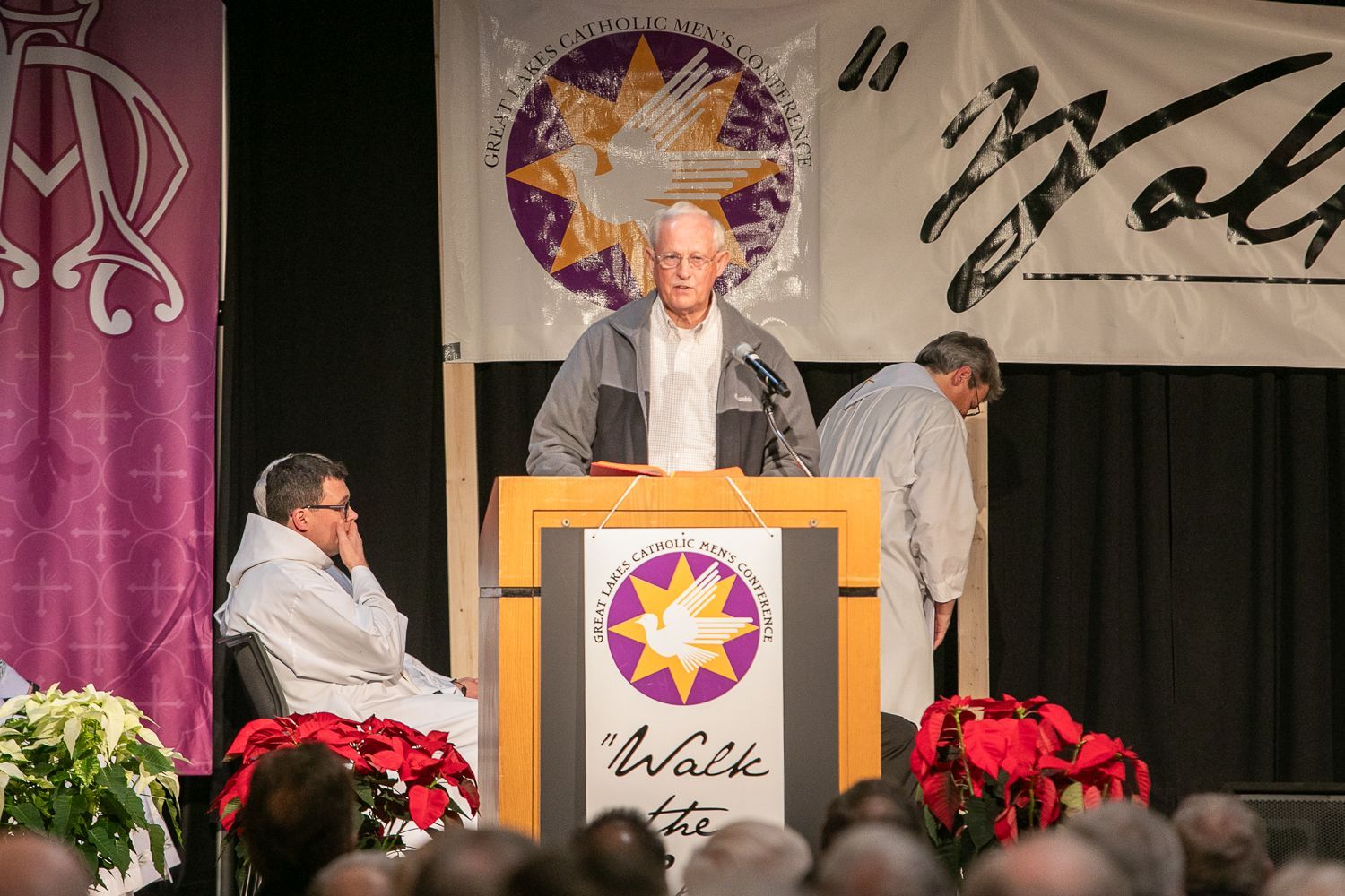 Speaker at podium on stage with purple and white banners, seated person at left, poinsettias in front
