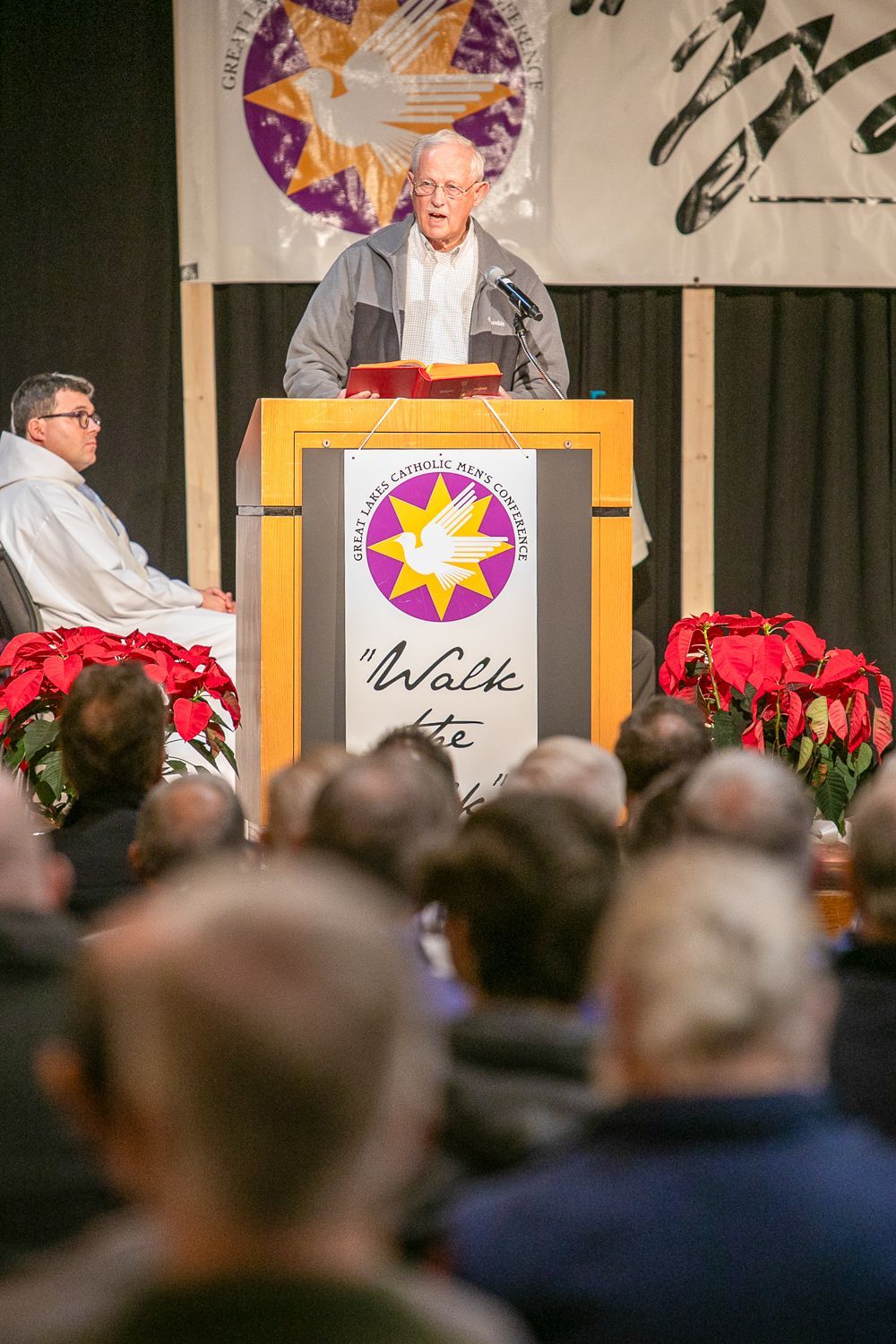 Man speaking at a podium on a stage, with audience in foreground and red flowers below.