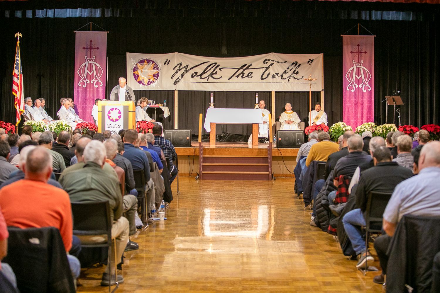 Audience seated in hall facing a stage with speakers, flowers, and a banner reading “Walk the Line”