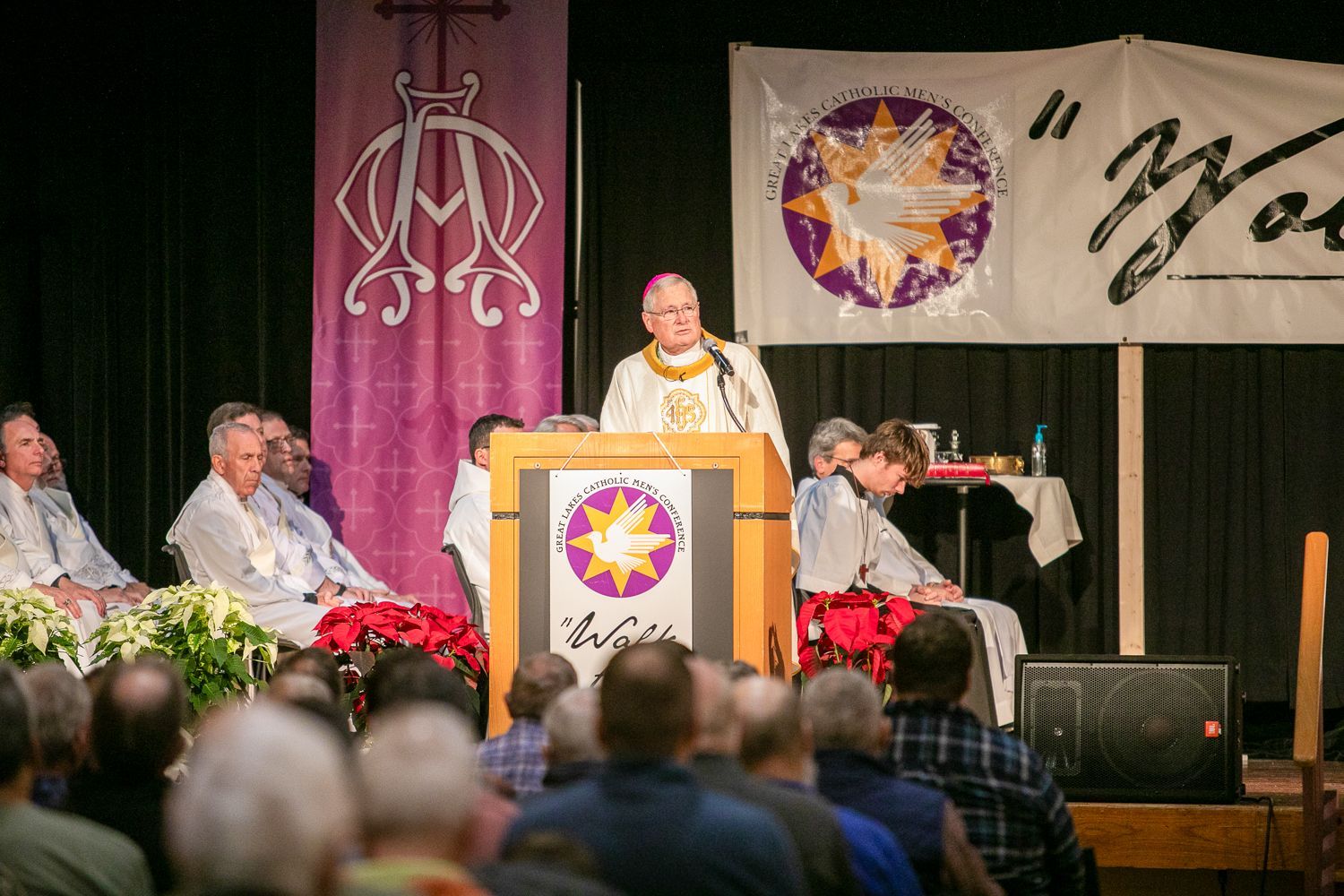 Bishop Malloy at podium on stage with banners, addressing an audience in an auditorium