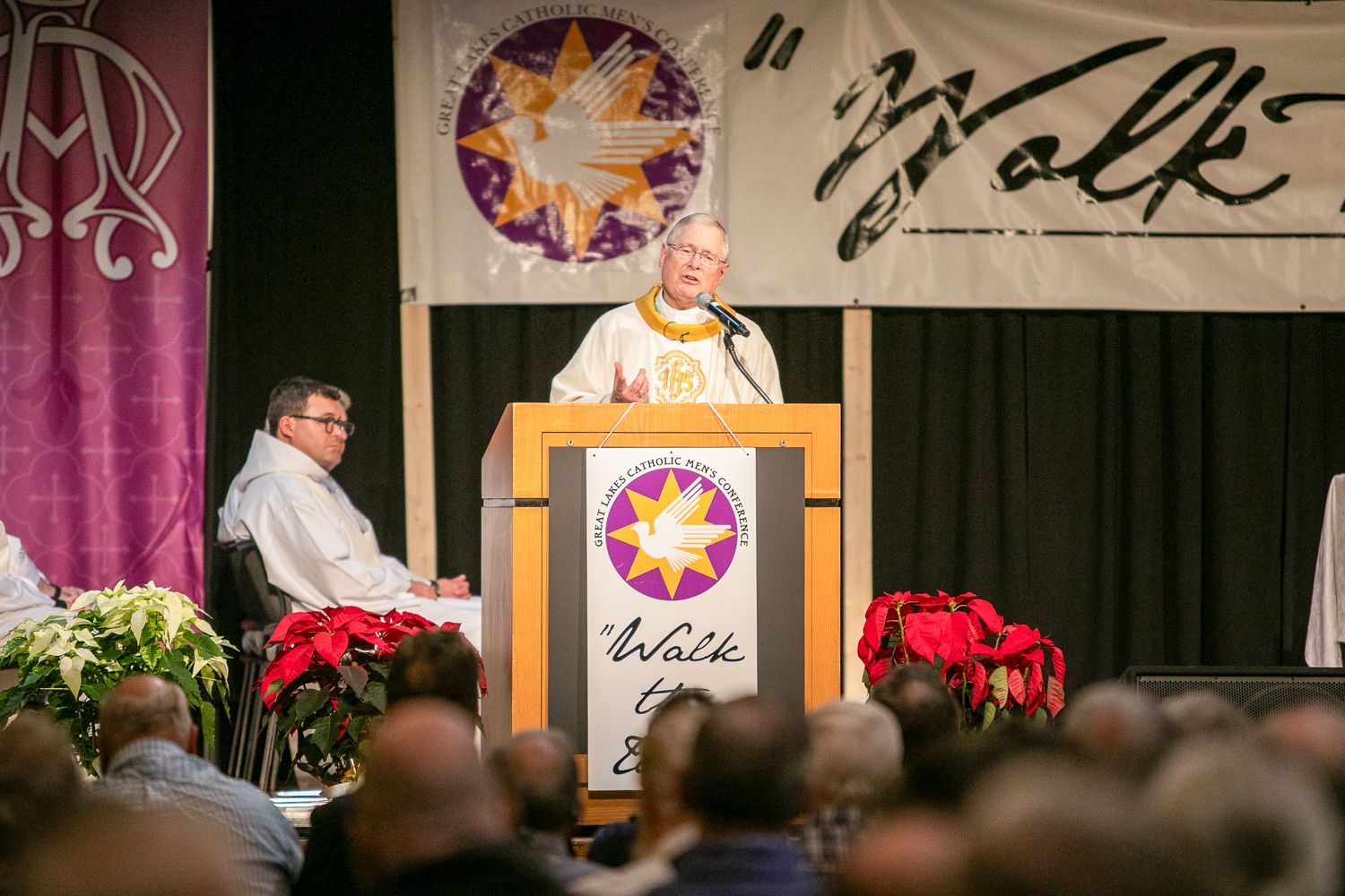 Bishop Malloy at a podium on a stage with a Wolf banner and red flowers, addressing an audience