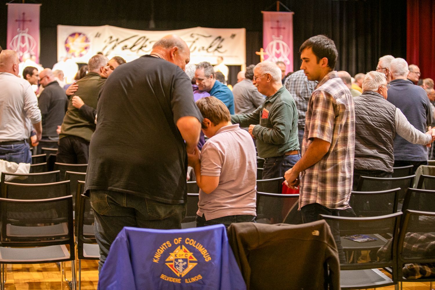 People standing and talking in a community hall during a gathering, with a stage and blue chair in the foreground.