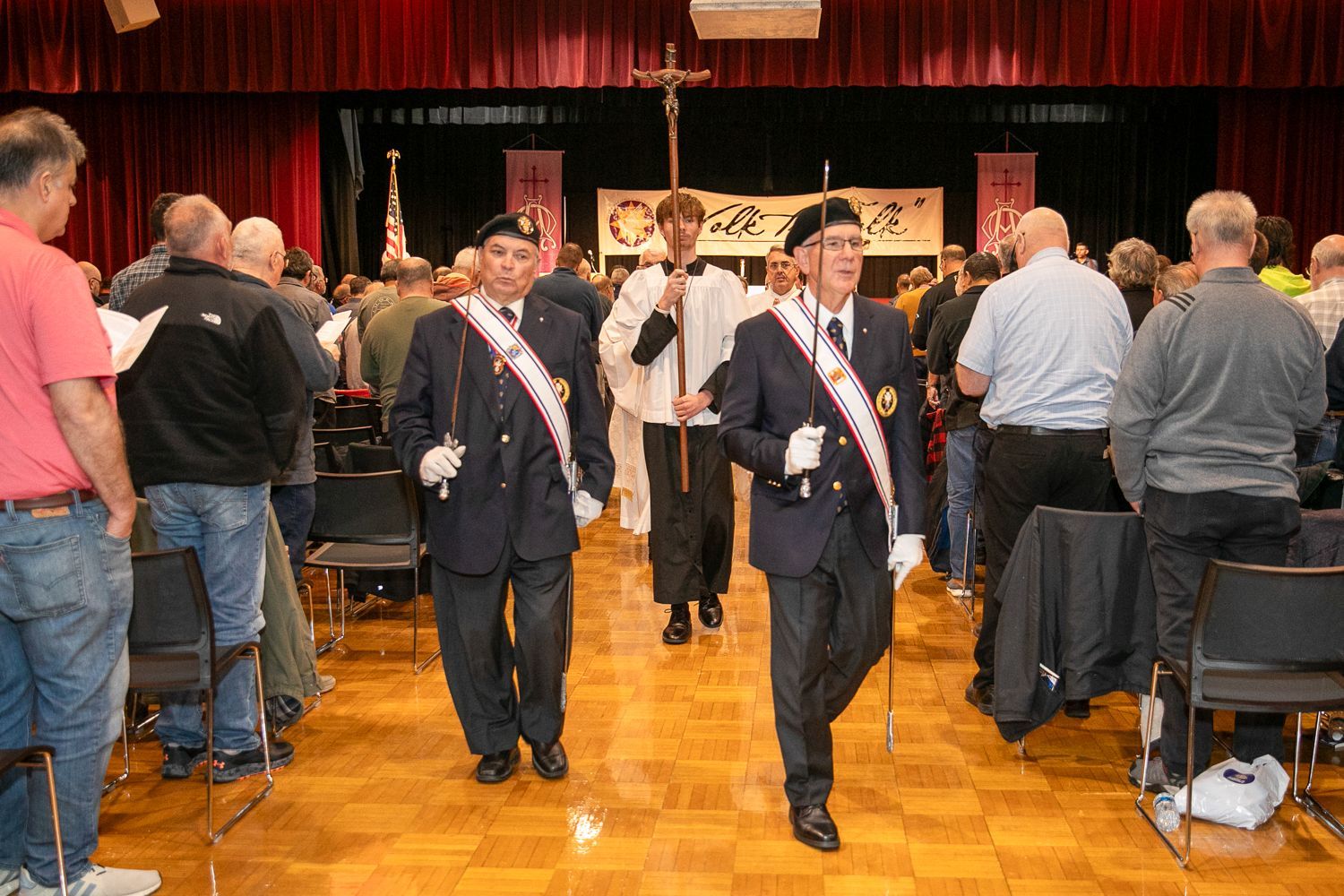 Two uniformed men march down an aisle in a crowded hall with people standing around them