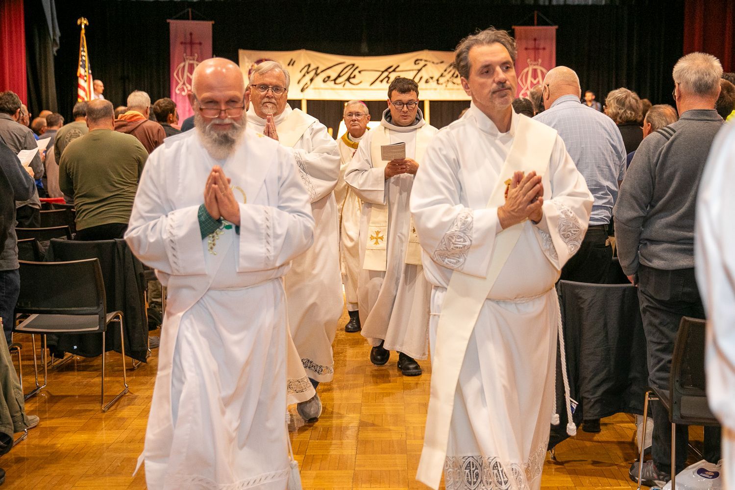 Two men in white robes walking in a church procession, hands clasped in prayer, with others behind them.