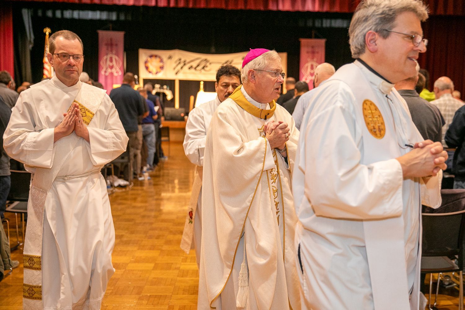 Clergy in white robes and a bishop in a pink zucchetto process through a banquet hall