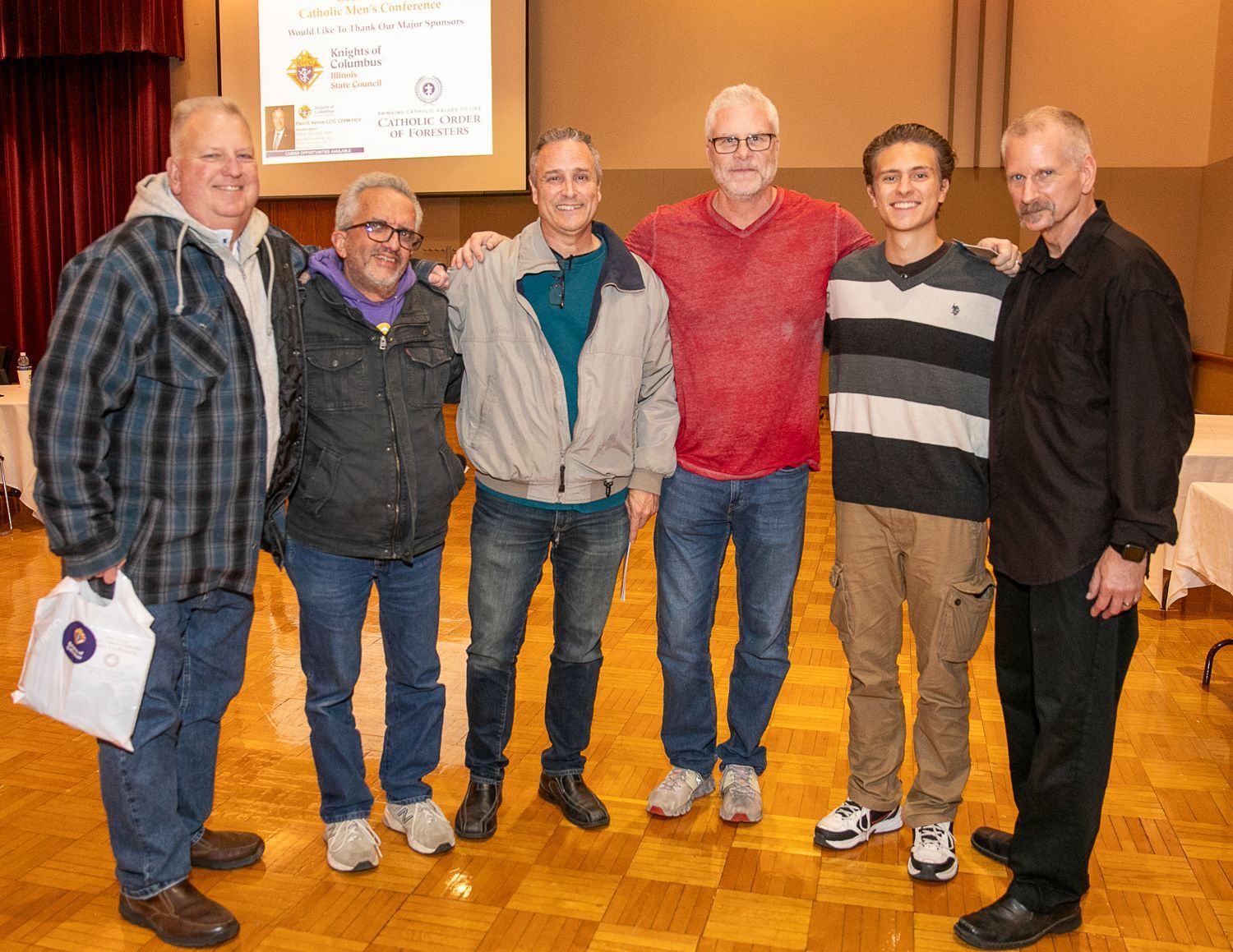 Six people stand together smiling in a wood-paneled hall with a stage in the background.