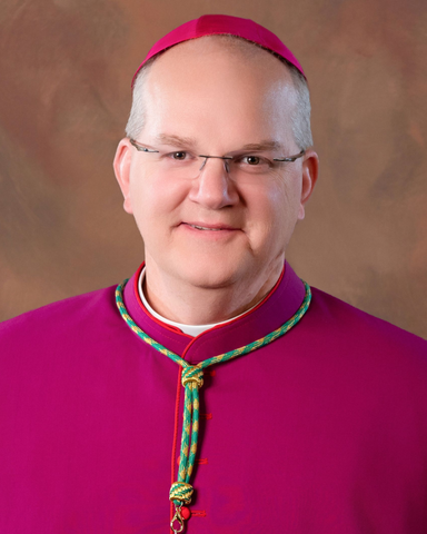Portrait of Fr. Rocky smiling in a dark suit and clerical collar against a warm studio background