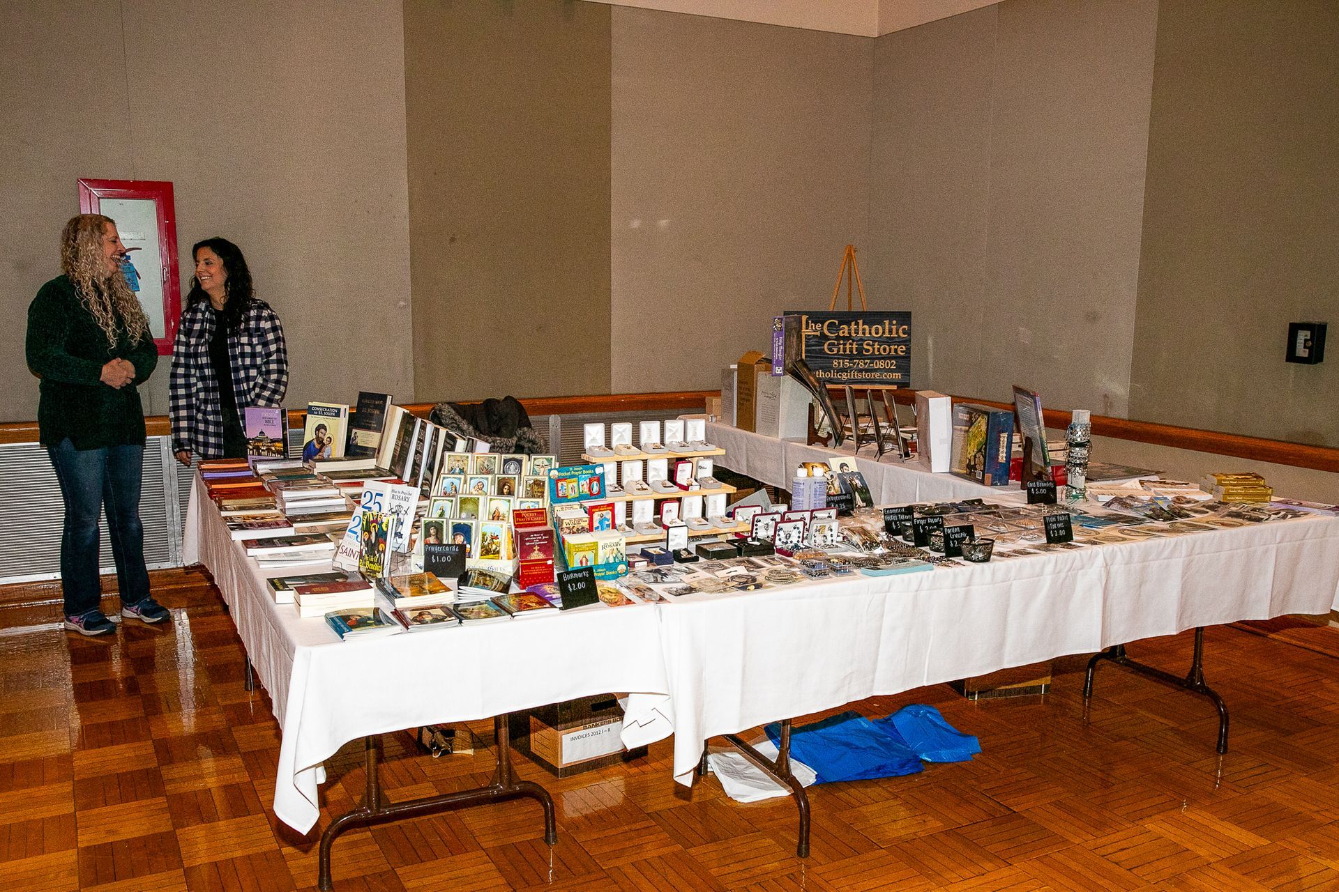 Tables of assorted items at a vendor booth, with two people standing in a hall.