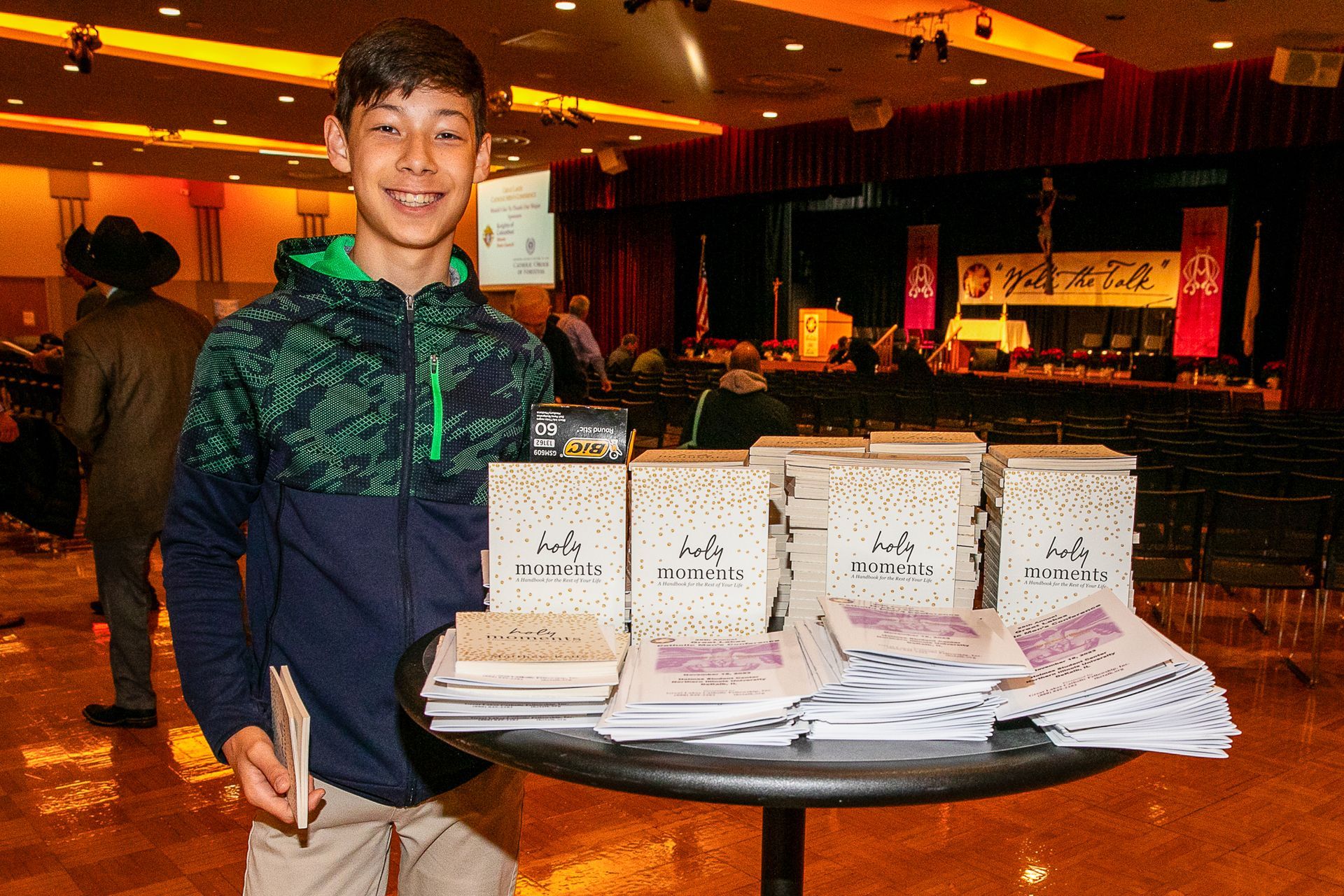 Person smiling behind a table of stacked books and pamphlets at an indoor event booth.