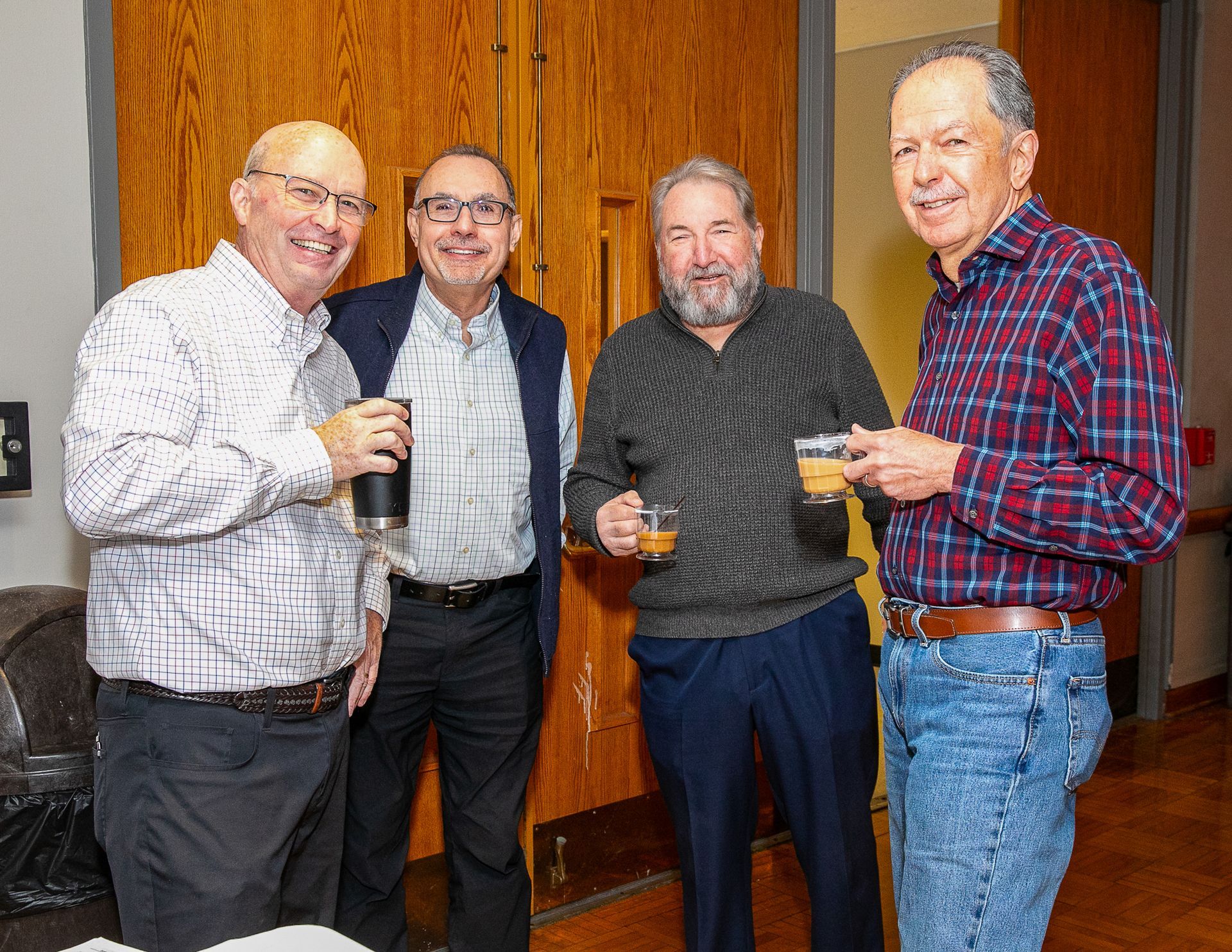 Four men standing indoors, smiling and holding drinks at a casual gathering.