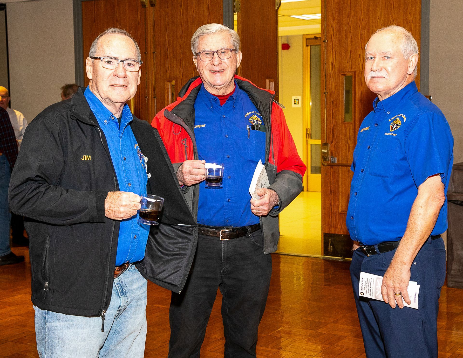Three men in blue shirts standing indoors, two holding drinks and one holding a pamphlet.