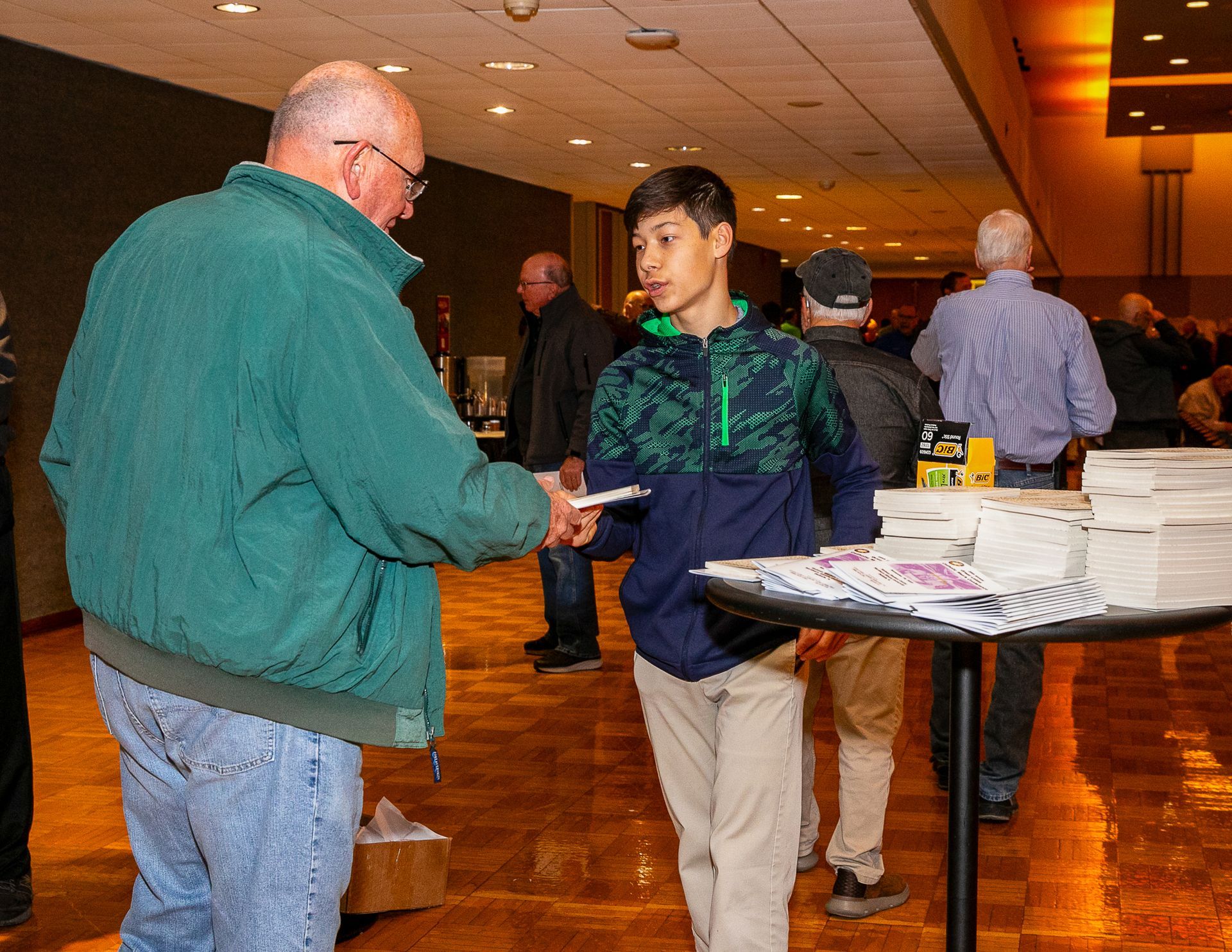 Two people talking at a table in a conference hall with stacks of papers nearby