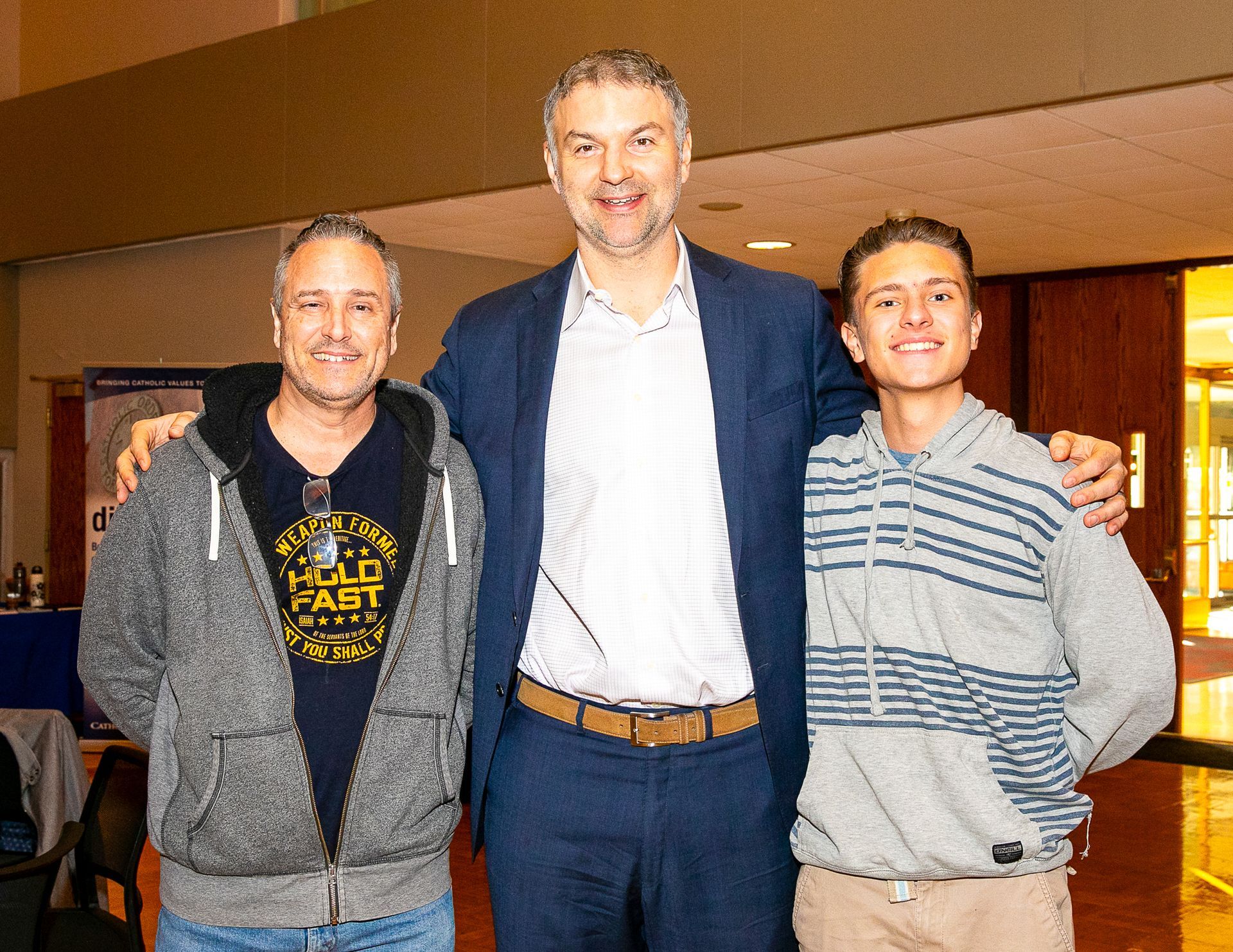 Three people smiling indoors, standing together with arms around each other.