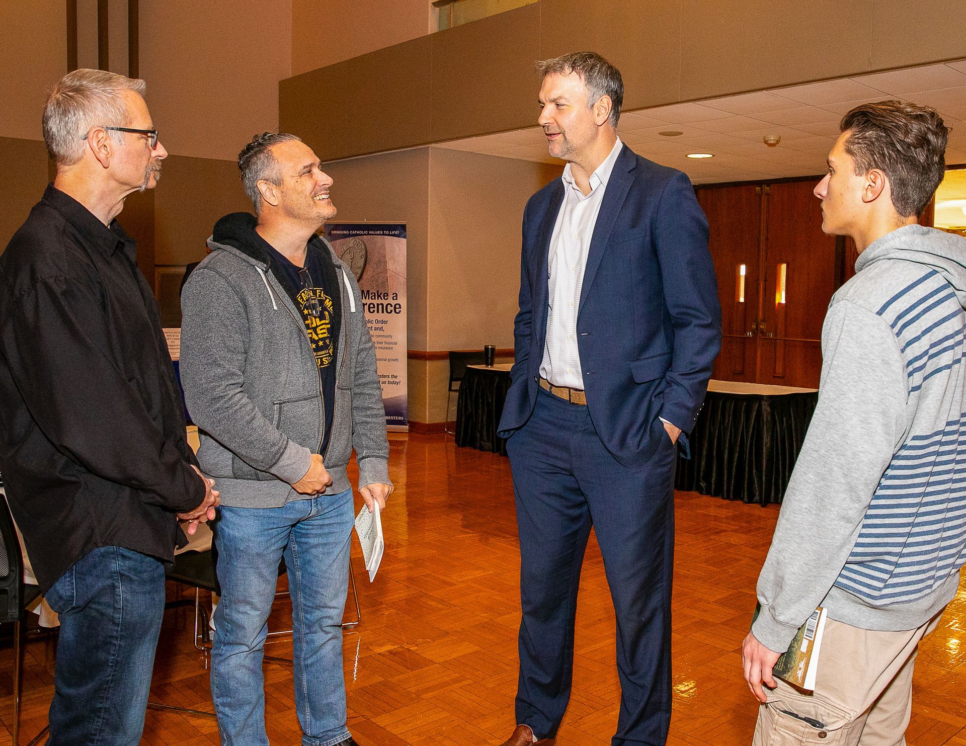 Four men chatting in a lobby, standing in a circle and smiling during a casual discussion.