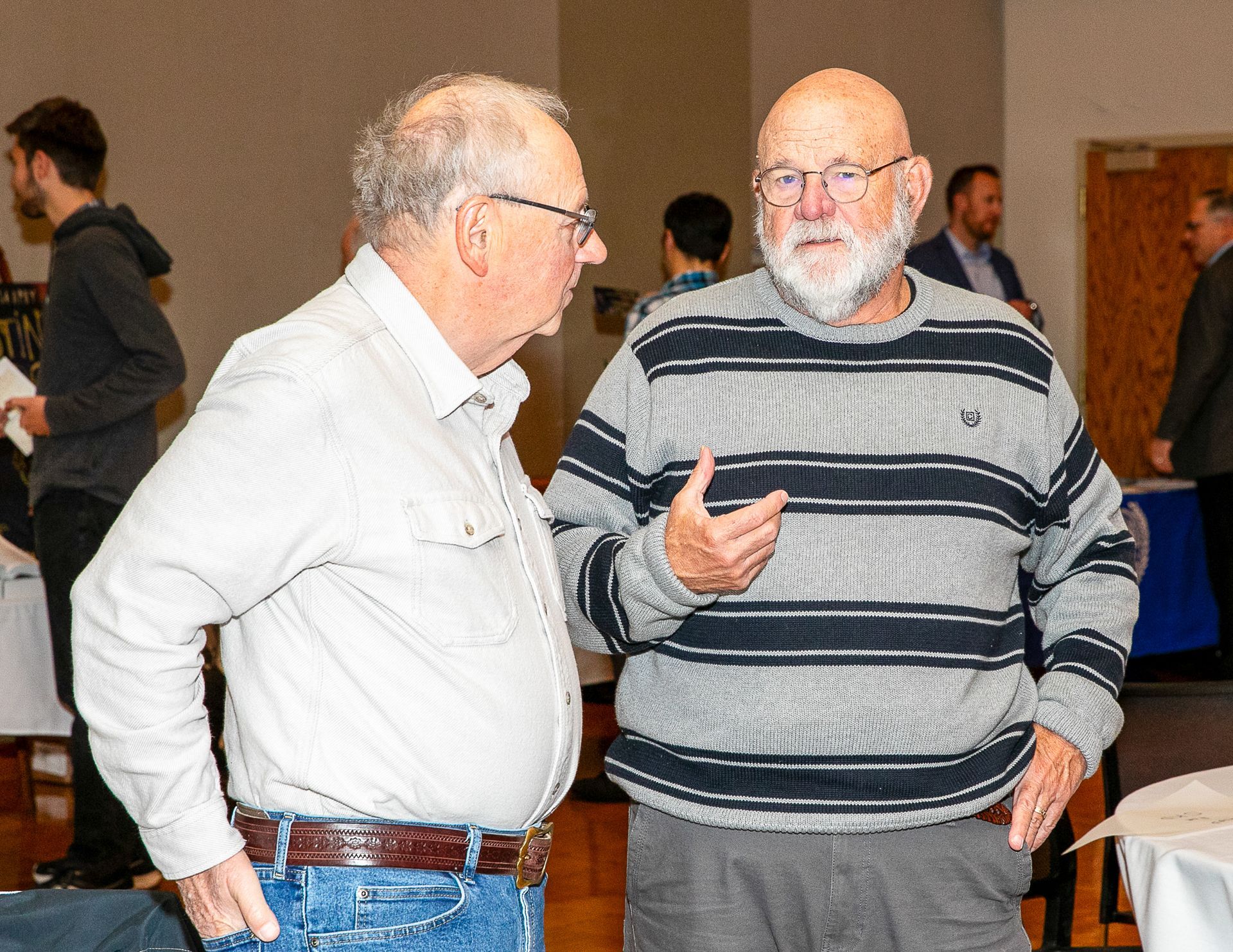 Two men talking indoors, one in a striped sweater gesturing, with others in the background.