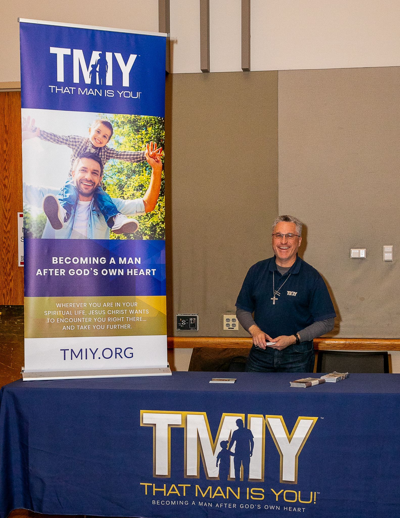 Man standing behind a TMY table banner at an indoor event with a matching tall poster nearby