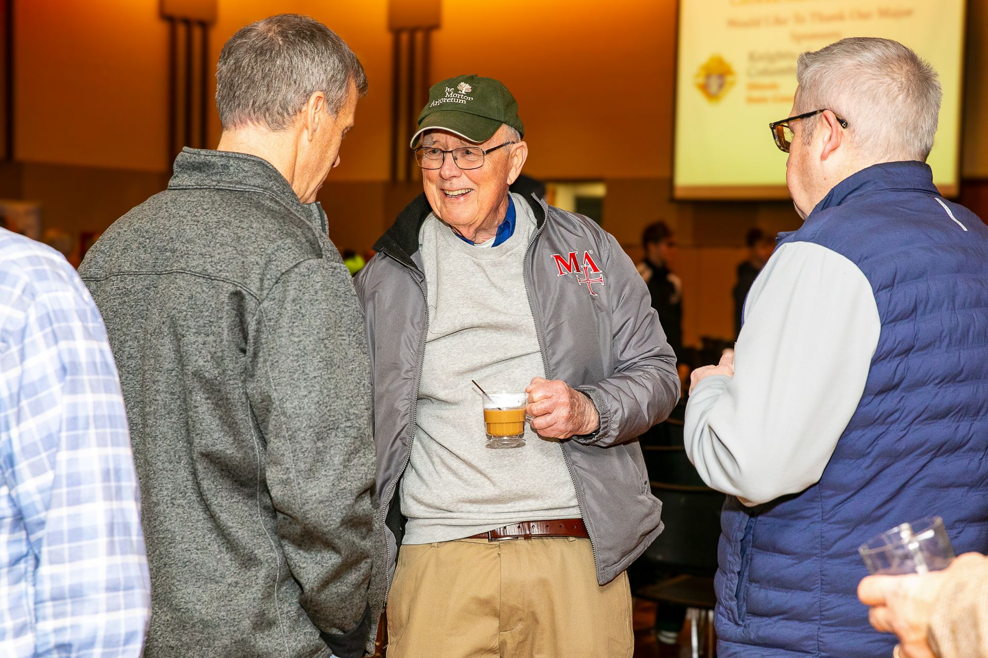 Three men chatting at an indoor event, one holding a drink, under warm lighting