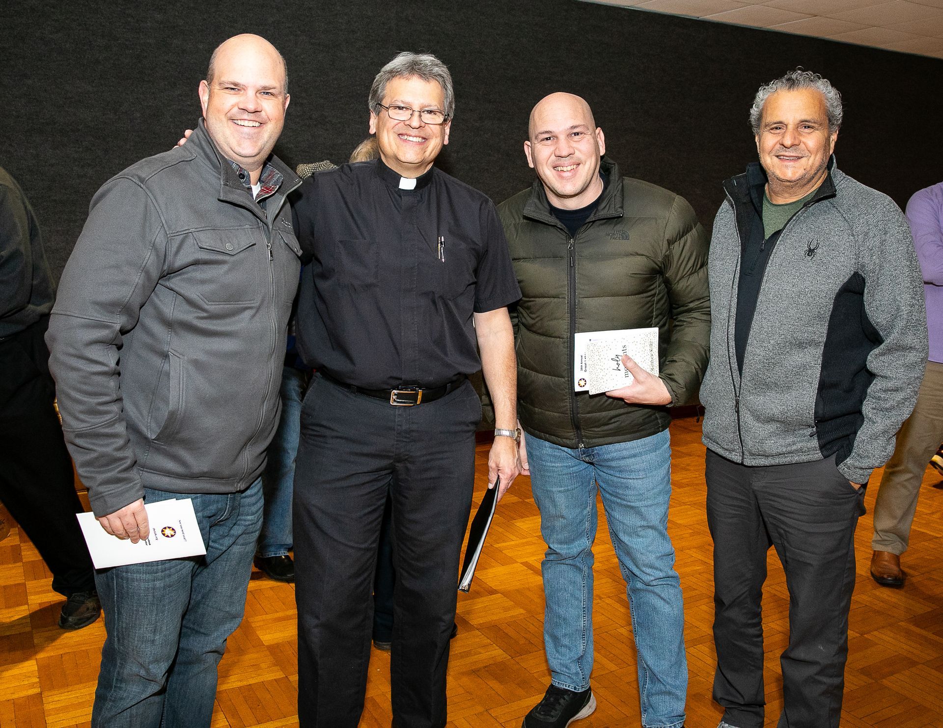 Four people standing indoors, smiling and holding small papers, on a wooden floor.