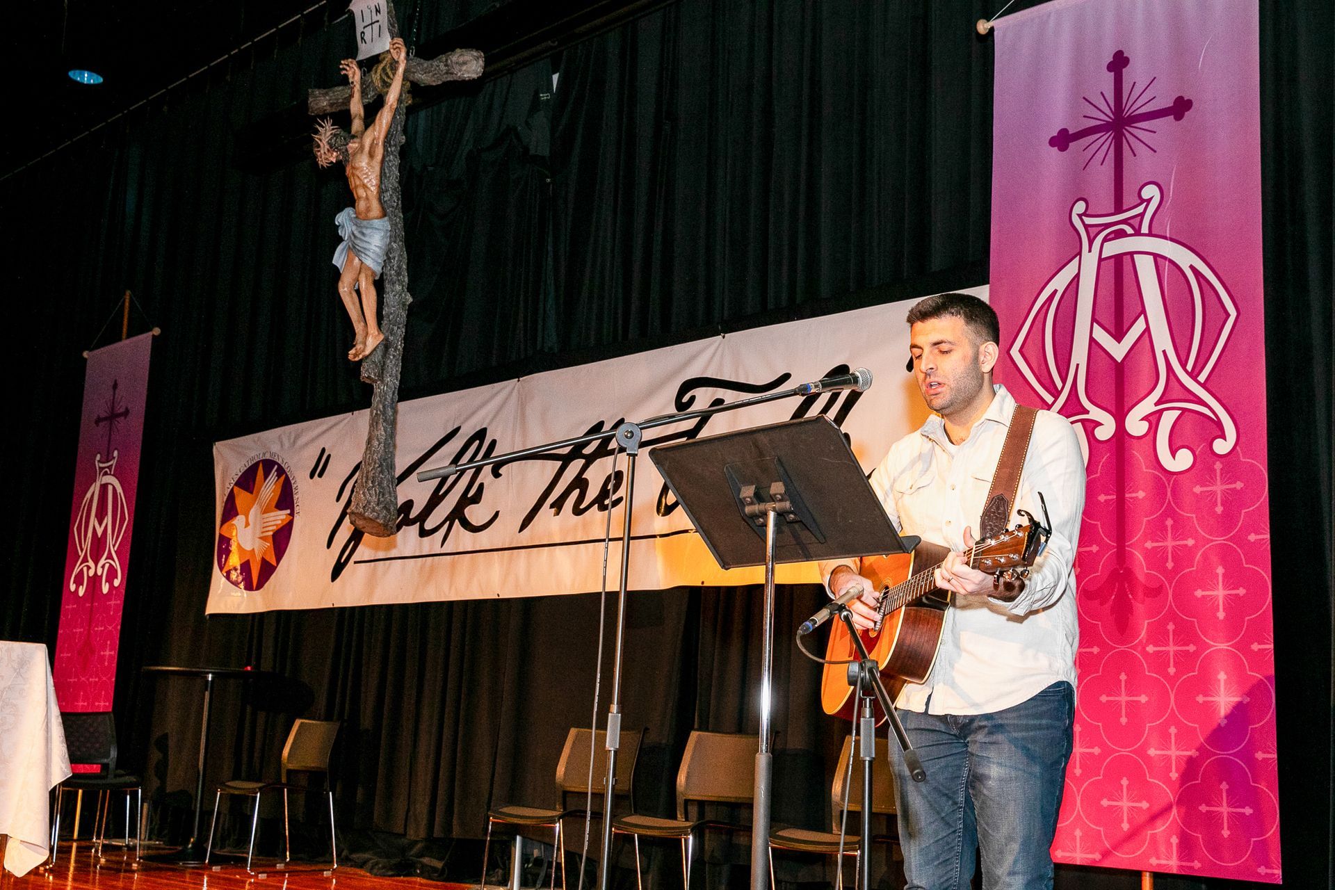 Musician playing guitar and singing on a church stage with crucifix and pink banners