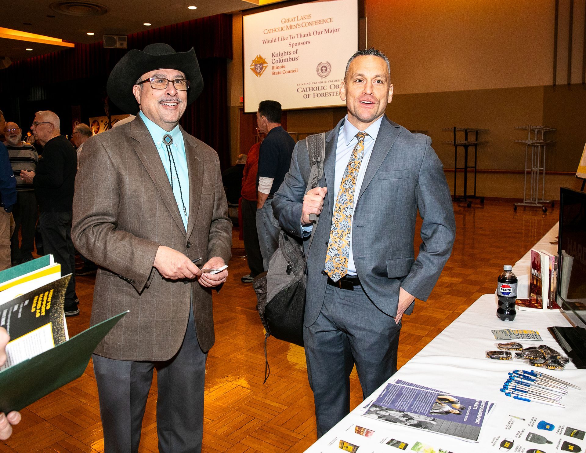 Two men in suits stand at a booth display in a conference hall.