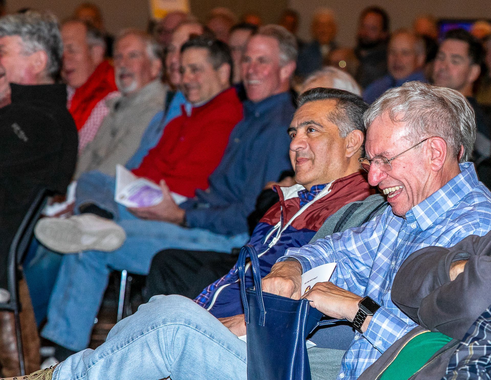 Audience seated in an auditorium, two men in front smiling and laughing during a presentation