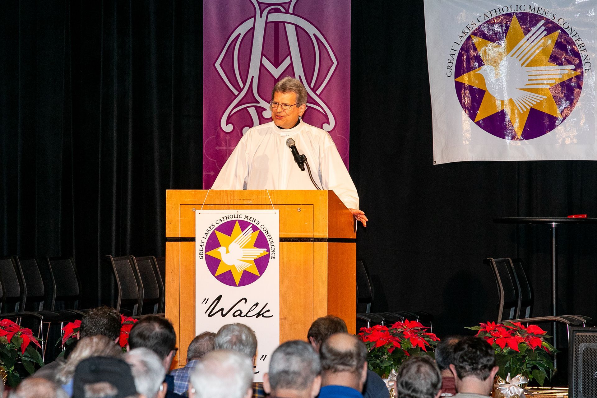 Speaker at podium on stage with purple and gold banner, addressing audience in an auditorium