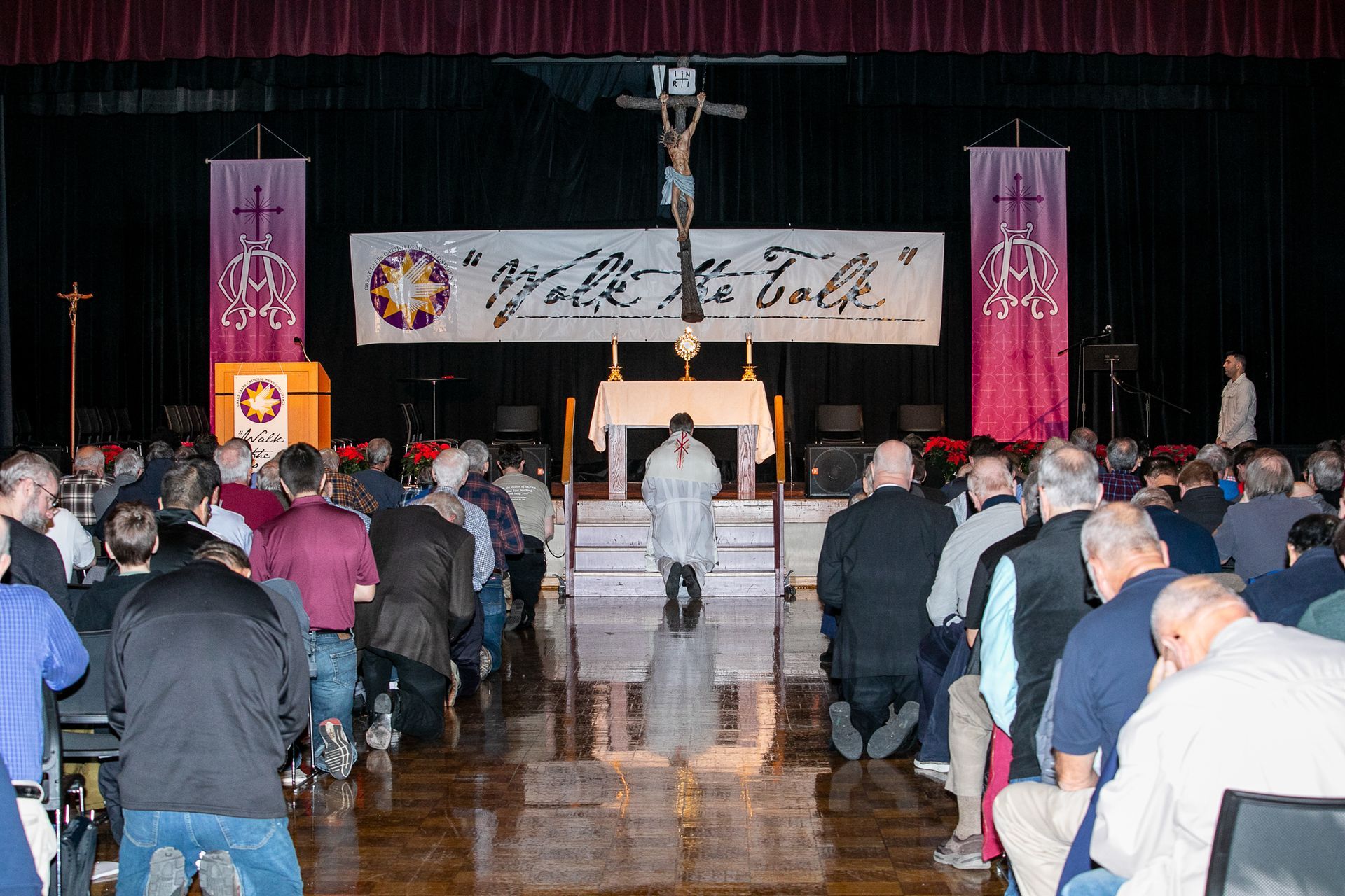 Audience facing a stage with a banner, podium, and altar in a large hall