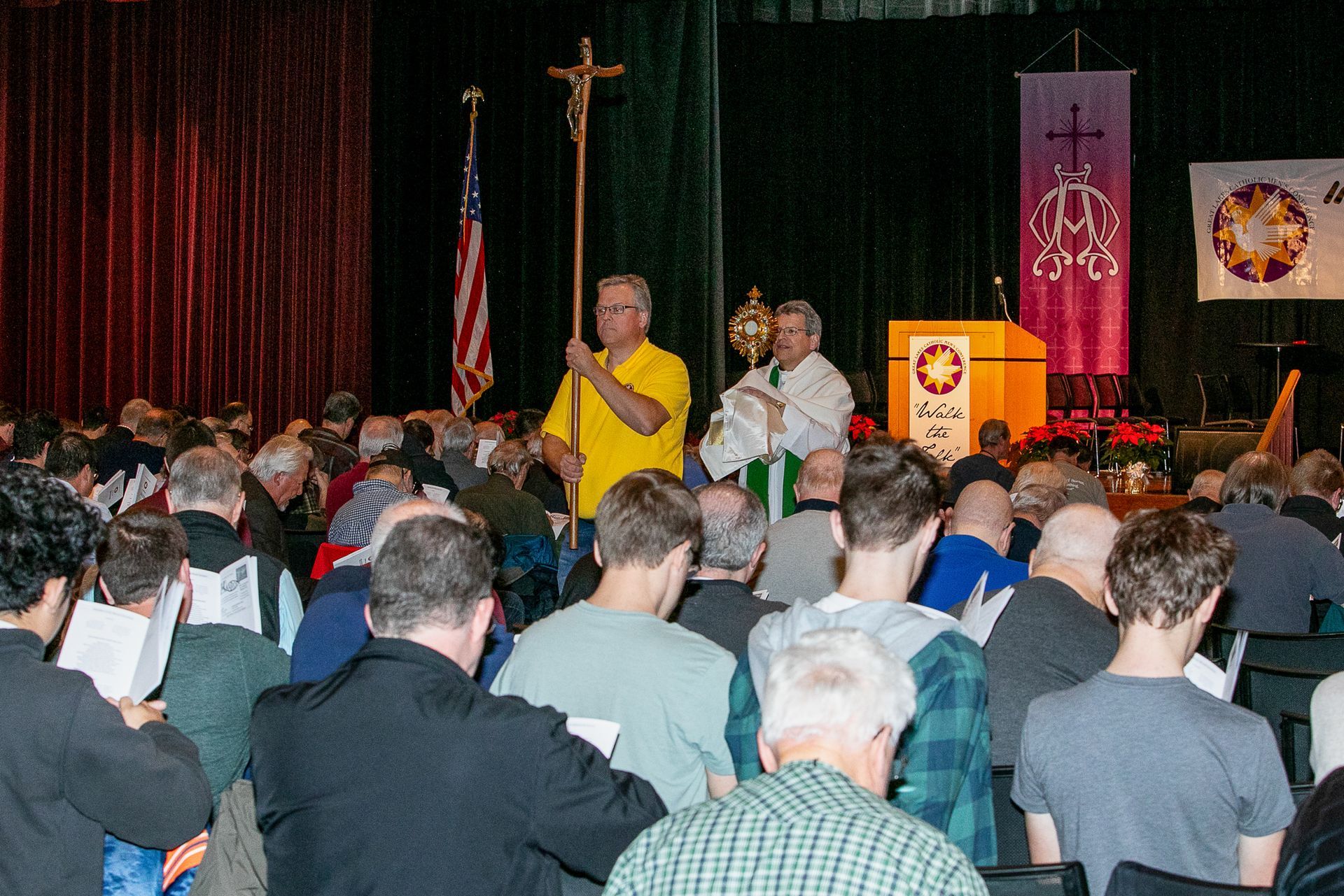 Audience seated facing speakers on a stage with a cross and religious banners, one speaker in yellow at a podium