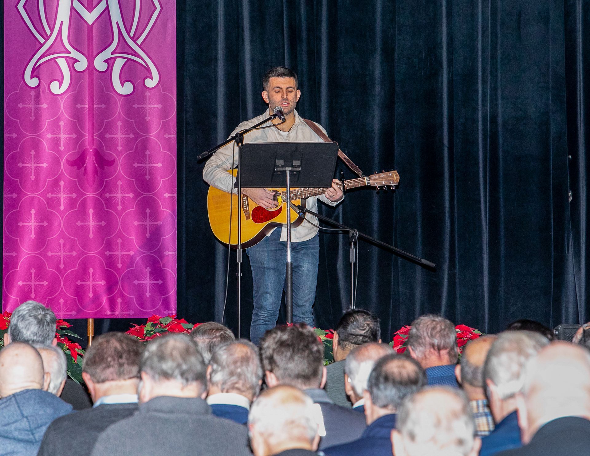 Musician sings and plays guitar on stage before an audience, with pink and dark blue curtains behind him