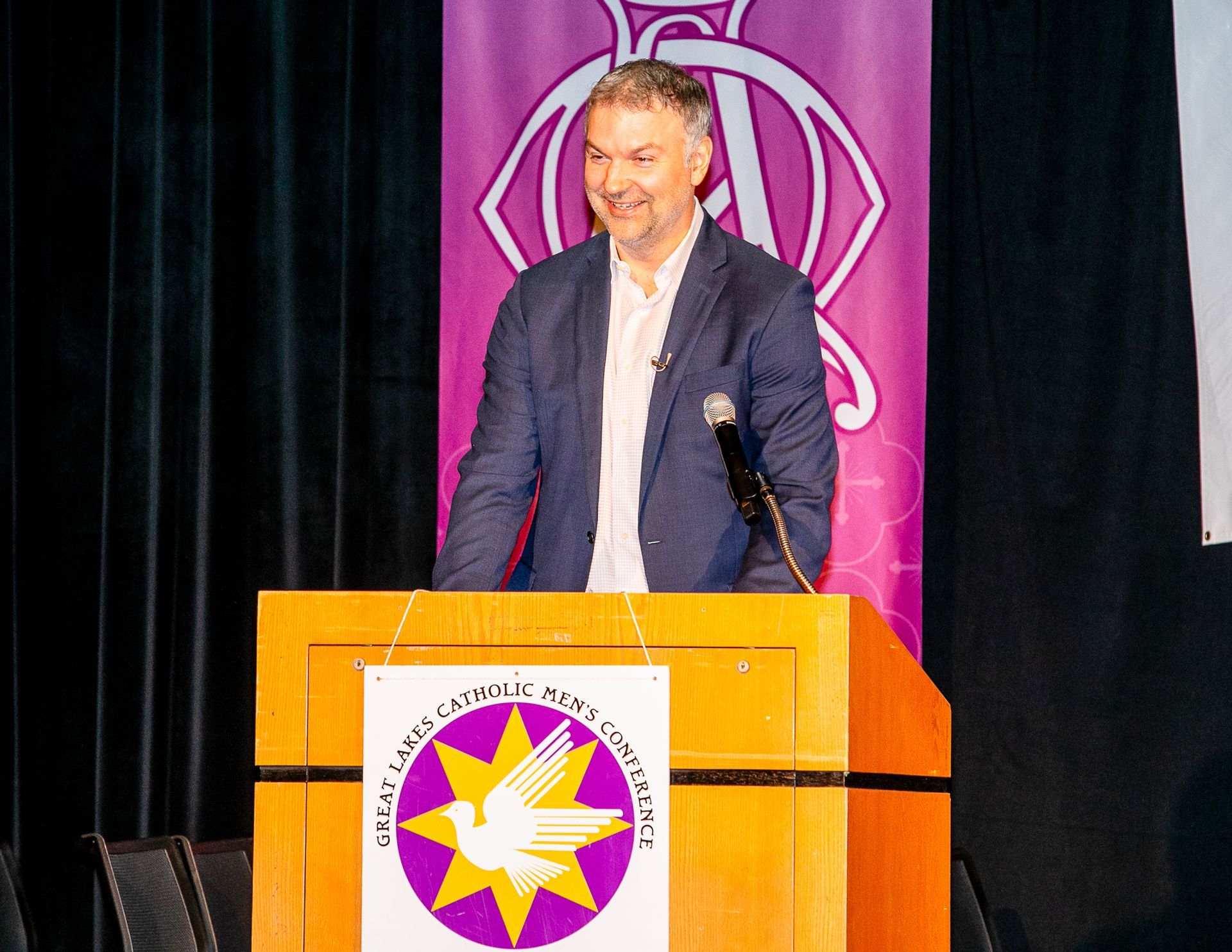 John Scott standing behind a podium on stage with a purple backdrop and emblem at a Catholic men's conference.