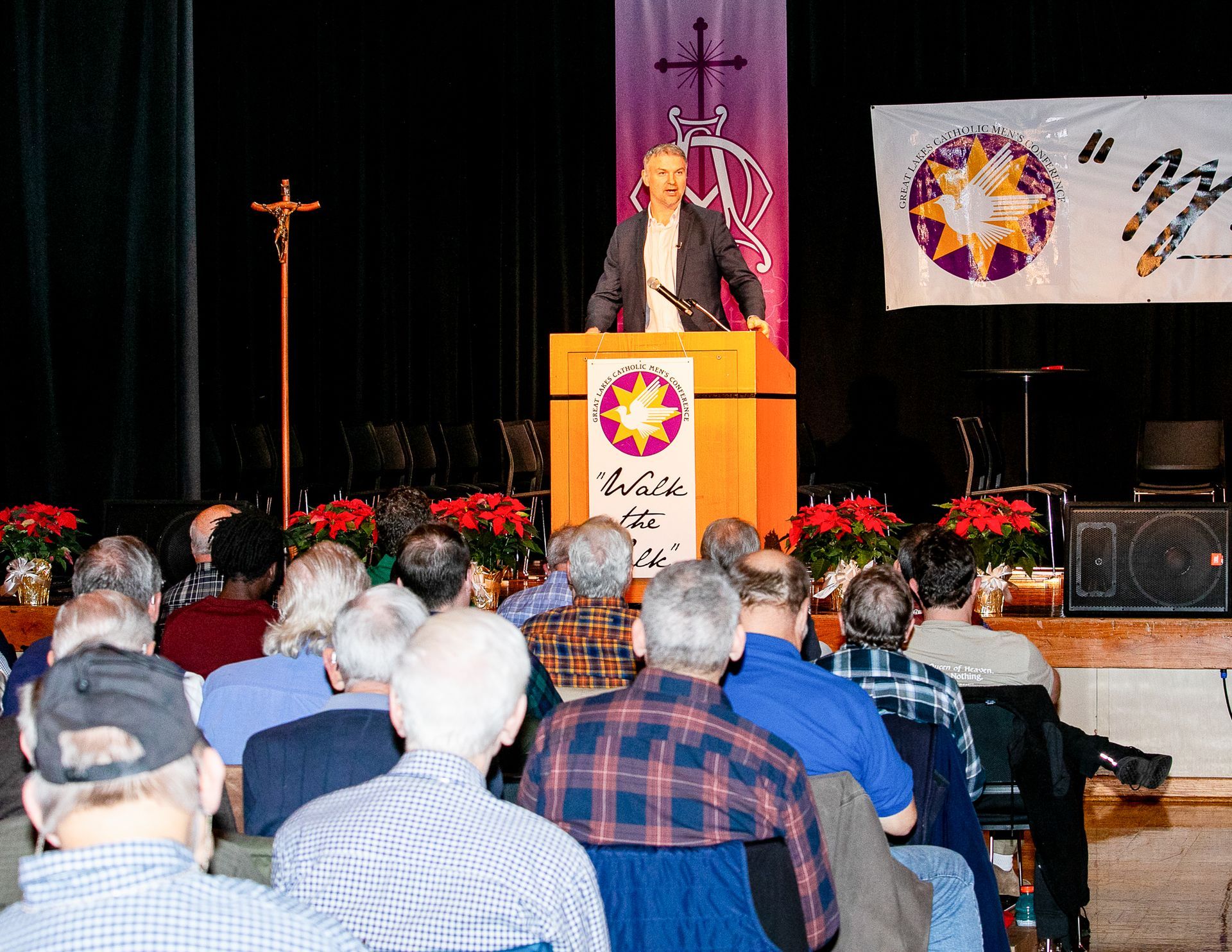 John Scott at podium addressing an audience in a decorated hall with stage lights and flowers