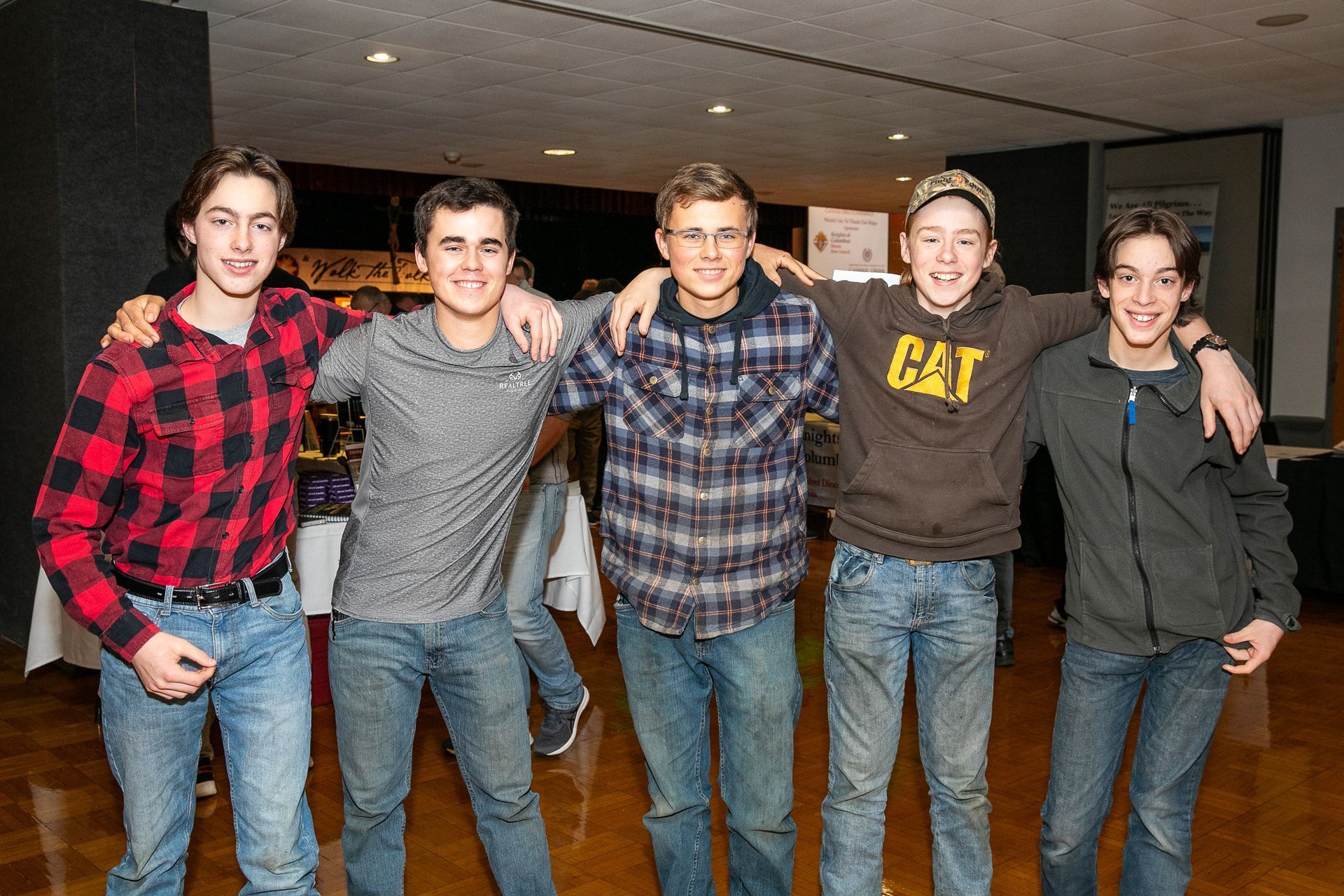 Five friends posing arm-in-arm indoors, smiling in casual plaid and jeans.