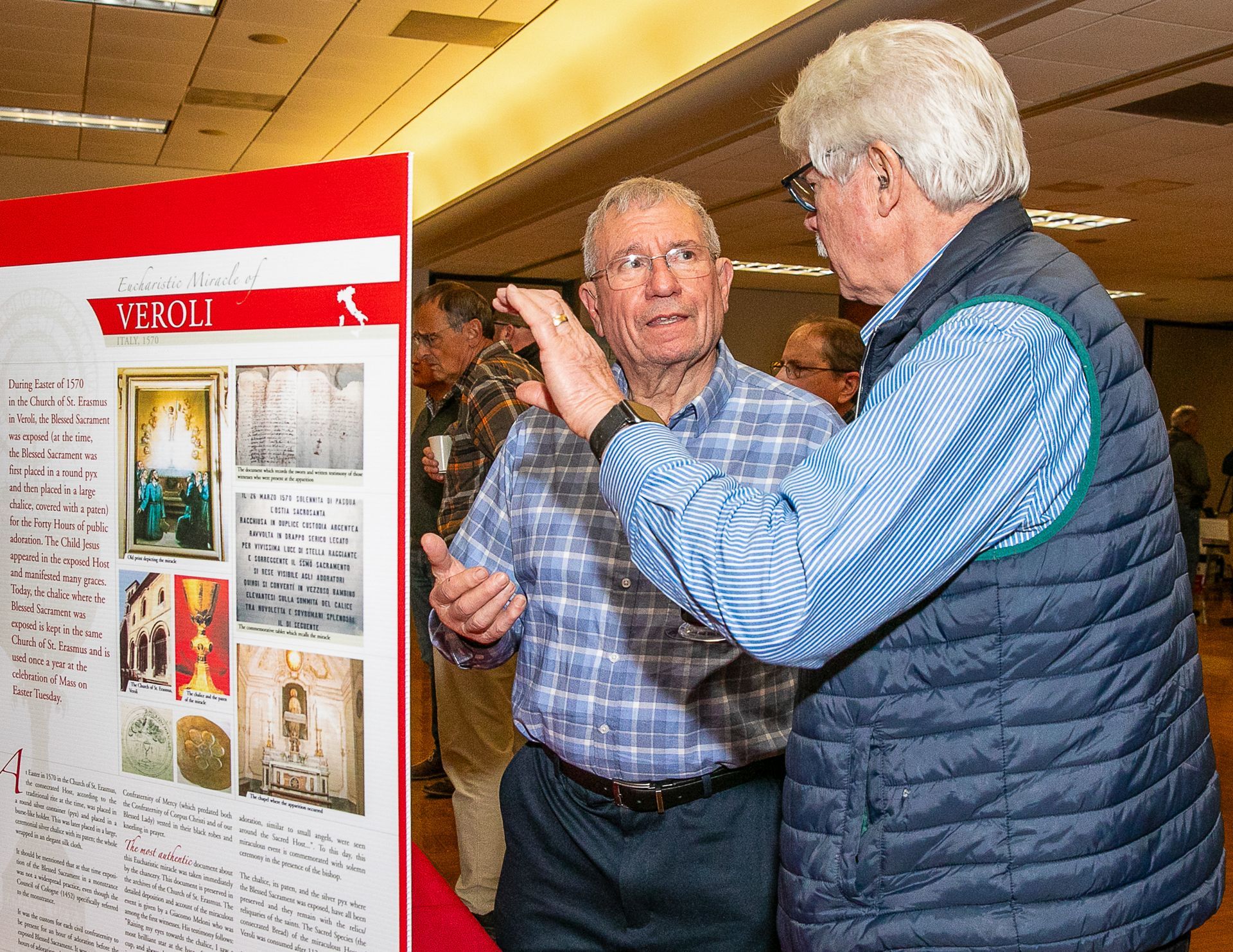 Two men talking beside an exhibit display board in a brightly lit indoor hall.